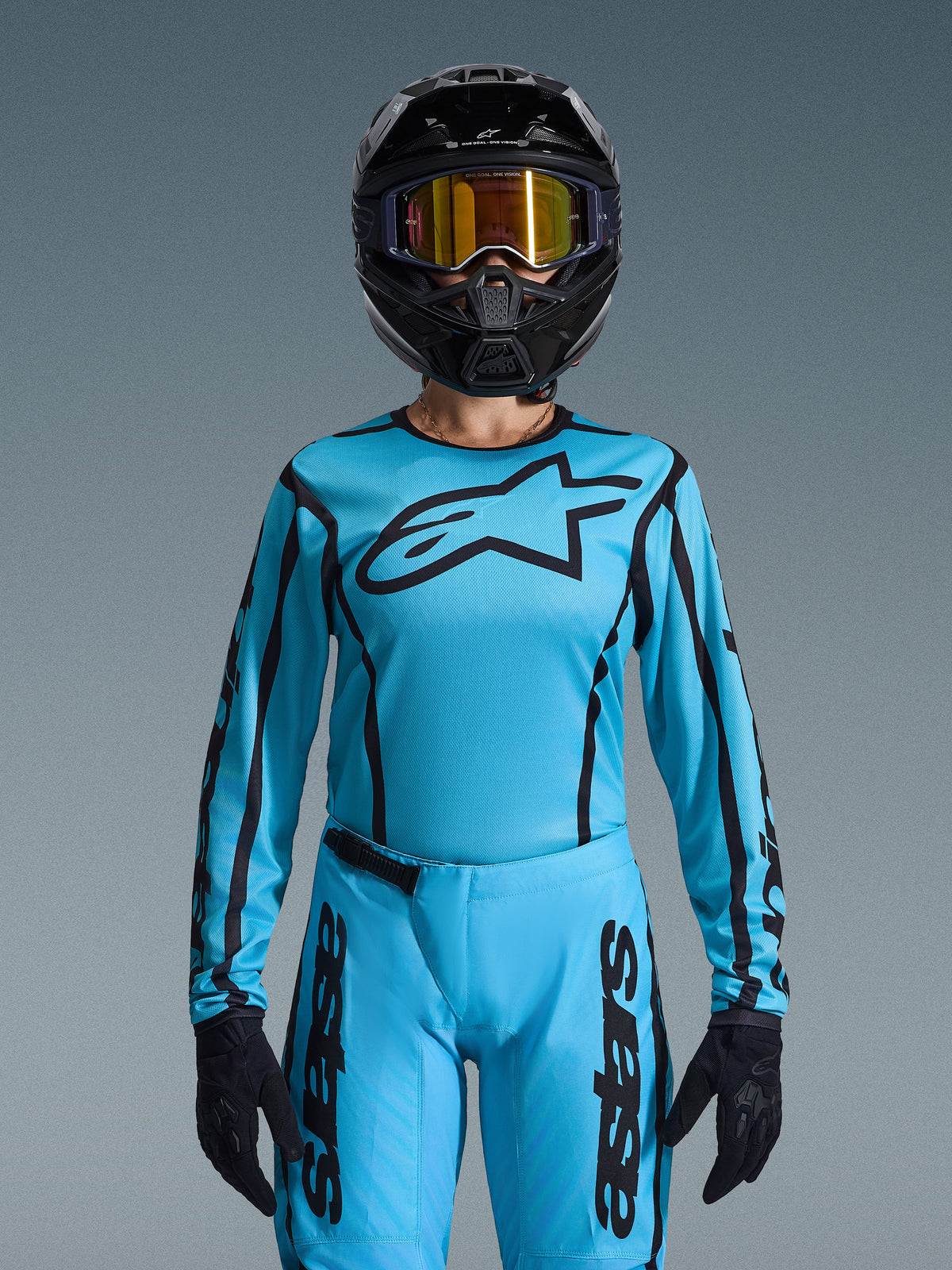 Piloto de motocross a usar o conjunto Alpinestars Stella Fluid Apex 2026 para mulher, camisola azul claro com contornos e logótipo pretos, calças a condizer, capacete preto com óculos iridescentes dourados e luvas pretas, conjunto de corrida de motocross feminino coordenado