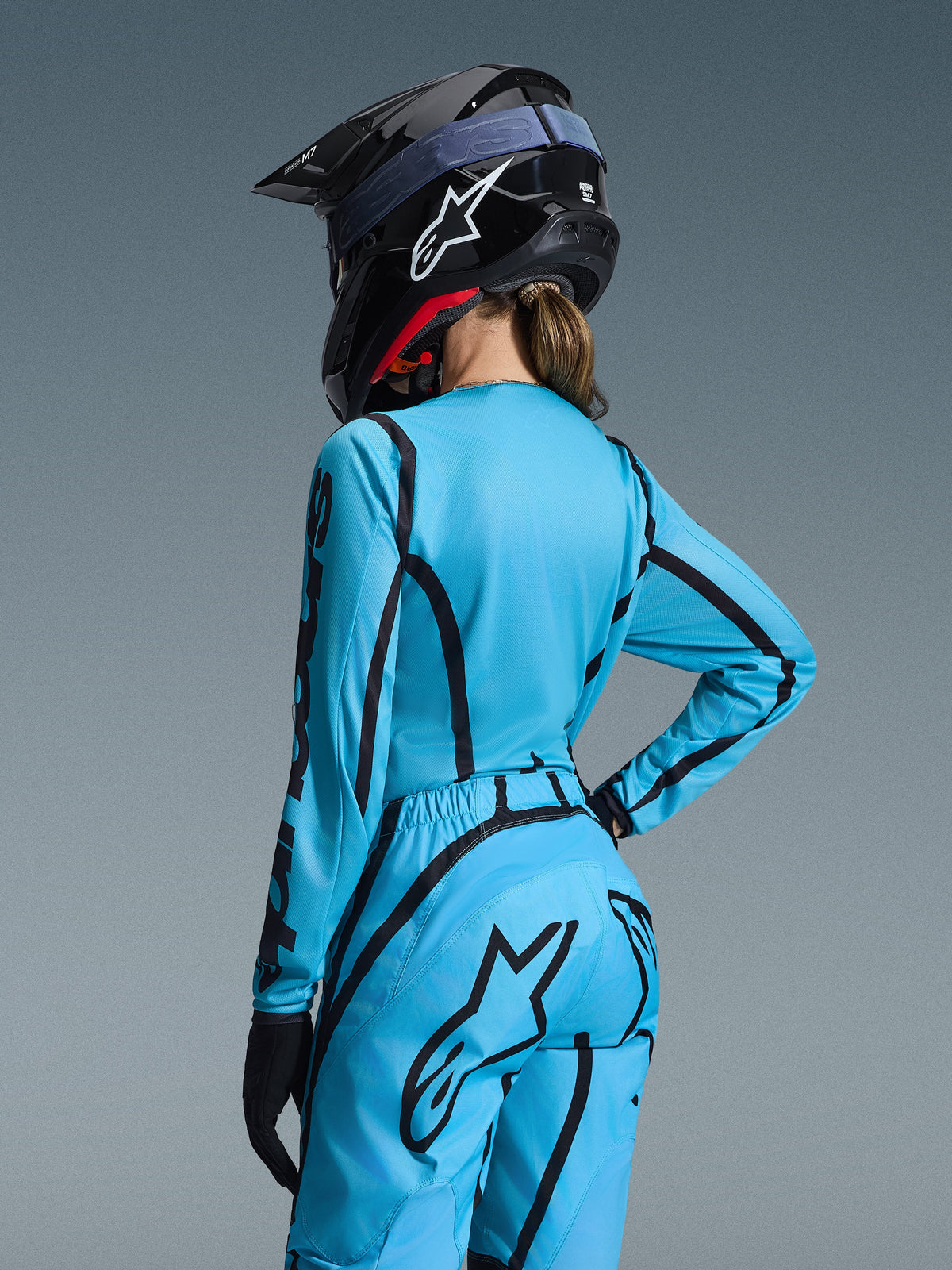 Conjunto de Equipamento Alpinestars Stella Fluid Apex 2026, azul claro e preto, vista traseira de uma piloto a usar camisola de motocross e calças a condizer, capacete Alpinestars preto com logótipo branco, vestuário técnico de corrida todo-o-terreno para mulher.
