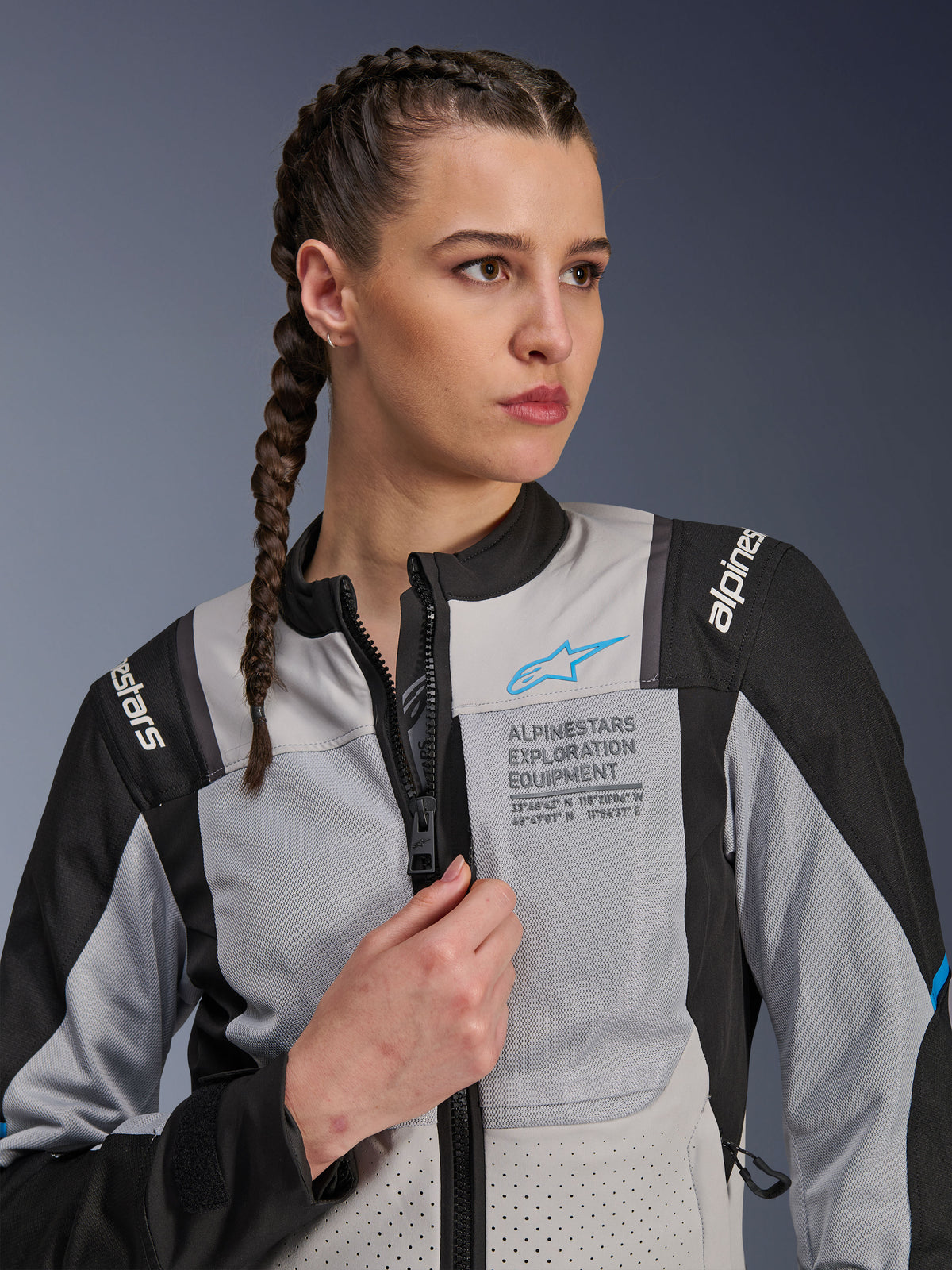 Mulher a usar o casaco Alpinestars Stella ST-2 Air em Ice Gray e preto, retrato em grande plano, com painéis de malha para ventilação, marca 'ALPINESTARS EXPLORATION EQUIPMENT' no peito com um logótipo azul, casaco de motociclismo de aventura moderno