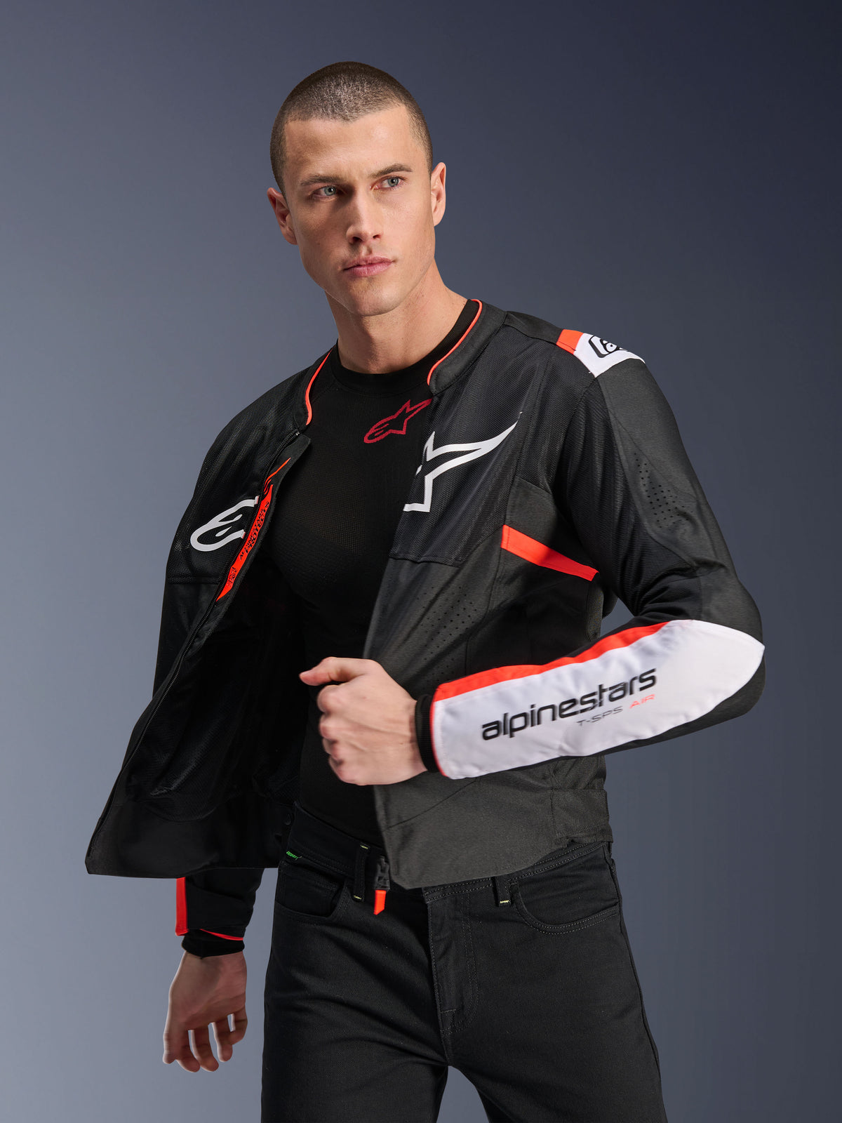 Modelo masculino a usar o Casaco Alpinestars T-SPS Air V2, casaco de motociclismo em têxtil preto com detalhes em branco e vermelho fluorescente, com painéis de malha para fluxo de ar, logótipo Alpinestars branco no peito e marca na manga, usado aberto sobre uma camada base preta
