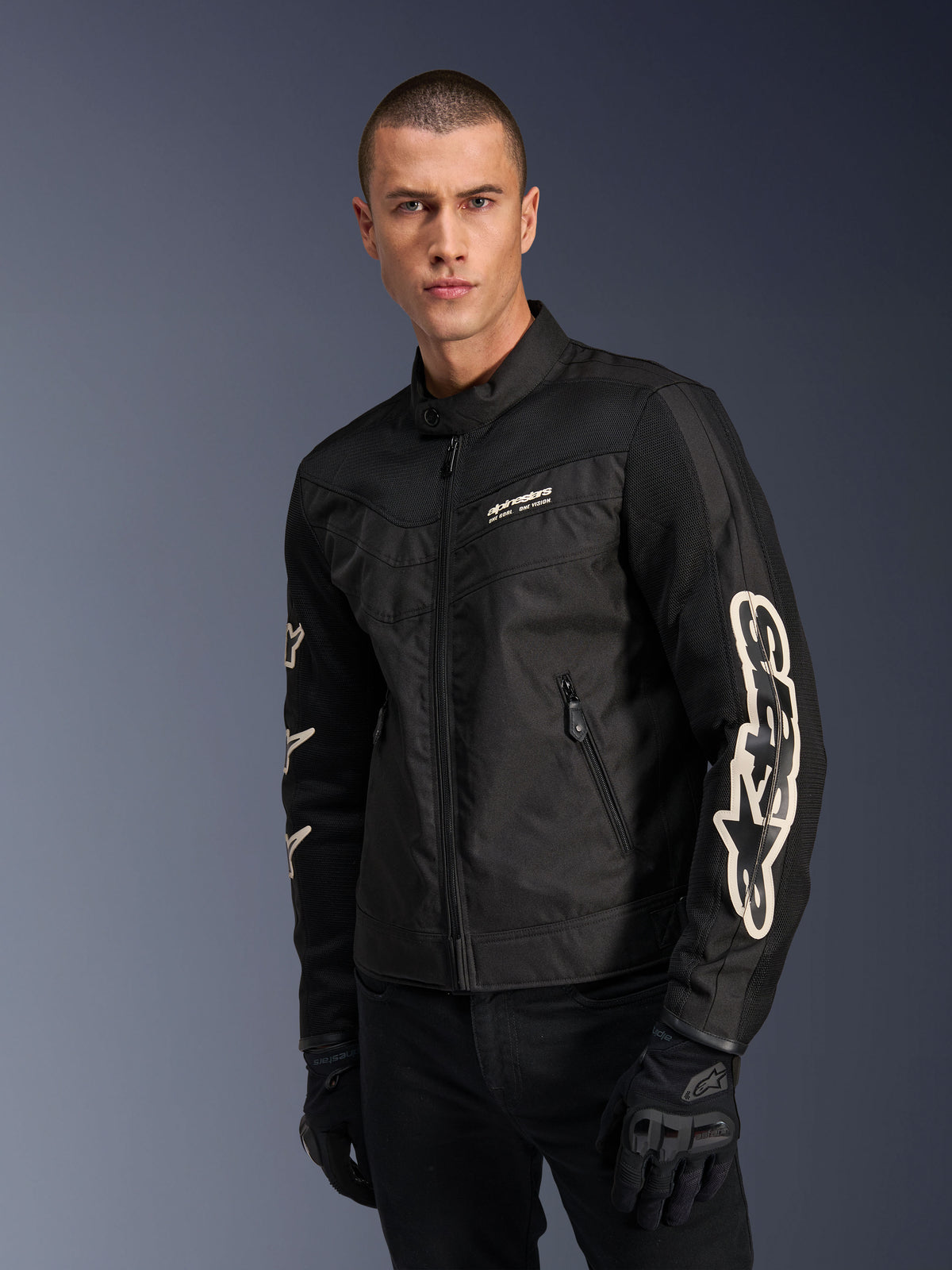 Casaco Alpinestars T-Dyno Air em preto, modelo masculino a usar um casaco de motociclismo têxtil de inspiração vintage com painéis de malha para ventilação, logótipo Alpinestars grande em branco nas mangas, acompanhado de luvas de condução pretas, design de equipamento de motociclismo urbano