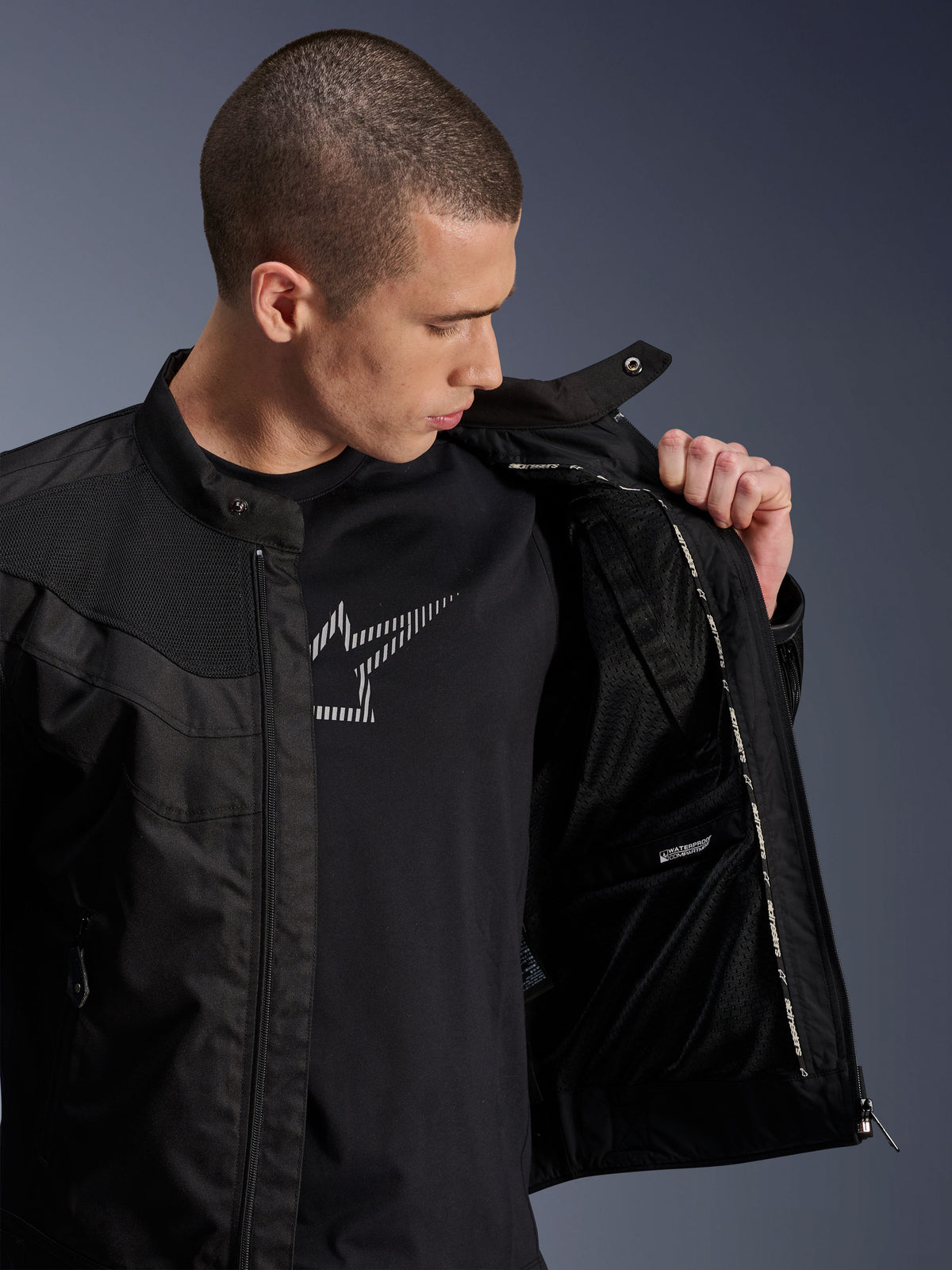 Casaco Alpinestars T-Dyno Air, preto, modelo masculino a mostrar o forro interno em malha e a etiqueta do compartimento impermeável, usado sobre uma t-shirt preta com grafismo, equipamento de motociclismo urbano moderno