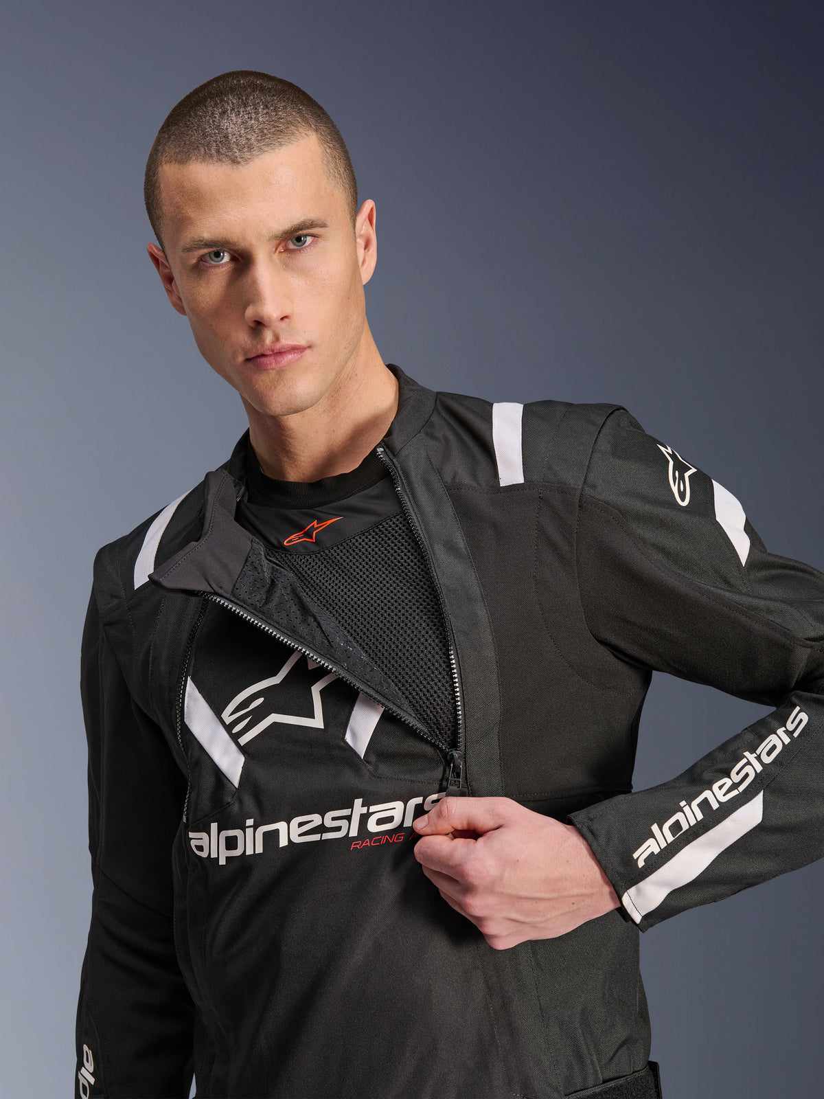 Alpinestars T-Stunt Air Jacket, casaco de motociclismo preto e branco, modelo masculino a mostrar o painel frontal modular parcialmente aberto revelando camadas de malha para fluxo de ar, logótipos Alpinestars brancos no peito e nas mangas, design desportivo para condução em estrada