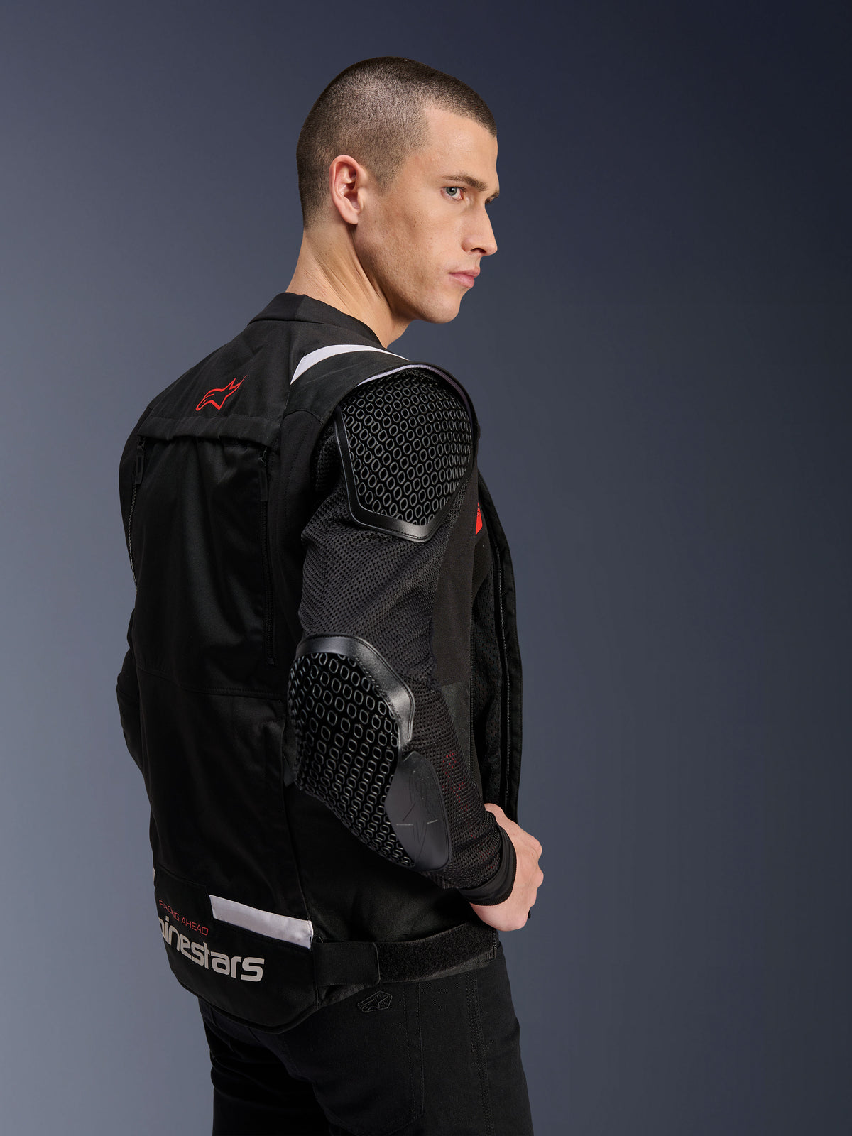 Casaco Alpinestars T-Stunt Air, casaco de motociclismo, preto, perfil lateral de um modelo a usar o casaco de corte desportivo com mangas de malha e proteção visível com padrão de favo de mel no ombro e cotovelo, logótipo vermelho nas costas, marca branca na cintura