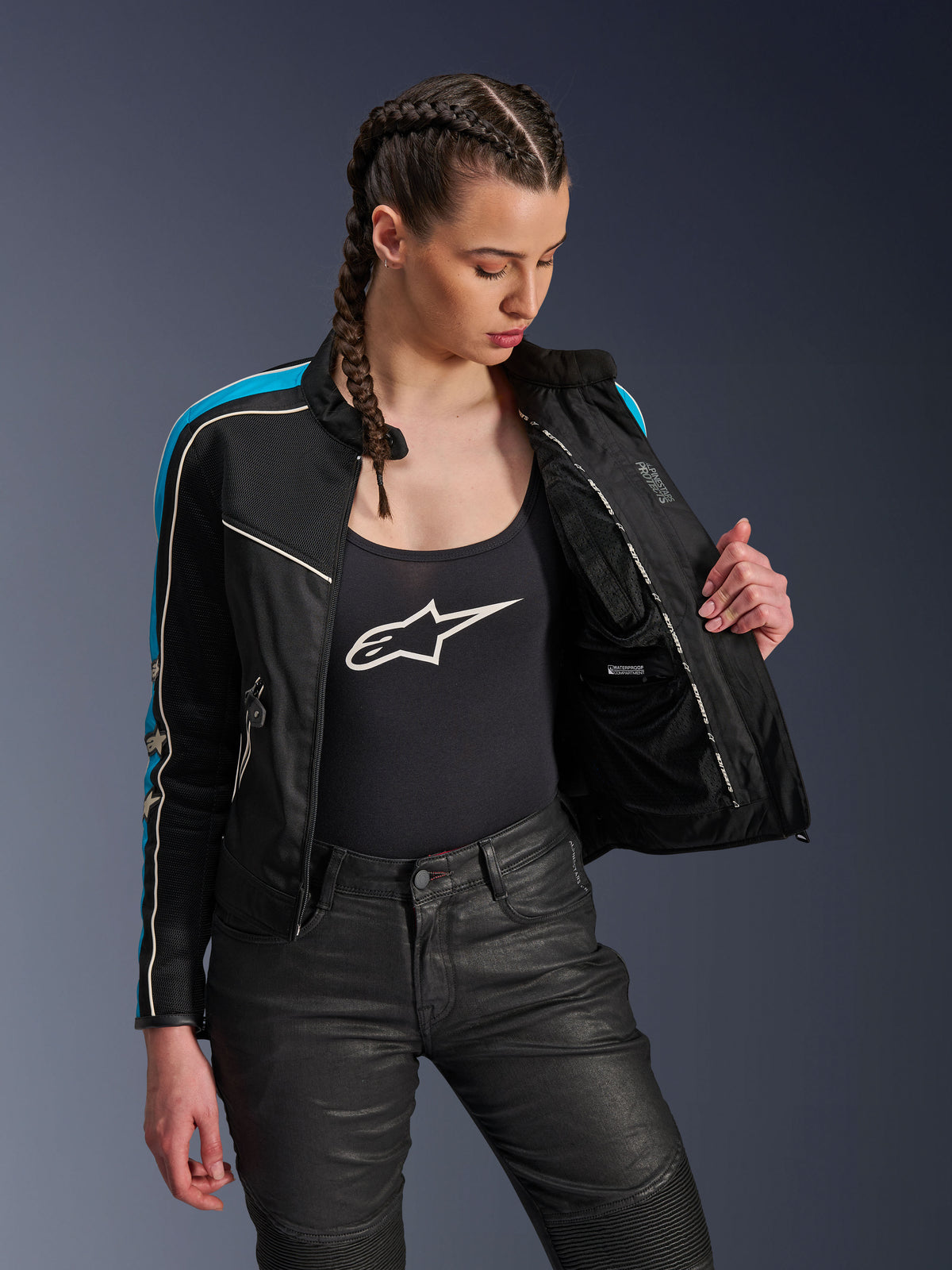 Mulher a usar o casaco Alpinestars Stella T-Dyno Air em Preto, Azul Claro e Ecru, segurando o casaco aberto para revelar o forro interno em malha e o bolso impermeável, combinado com um top de alças preto com logótipo e calças de condução pretas, equipamento de motociclismo urbano de inspiração vintage.