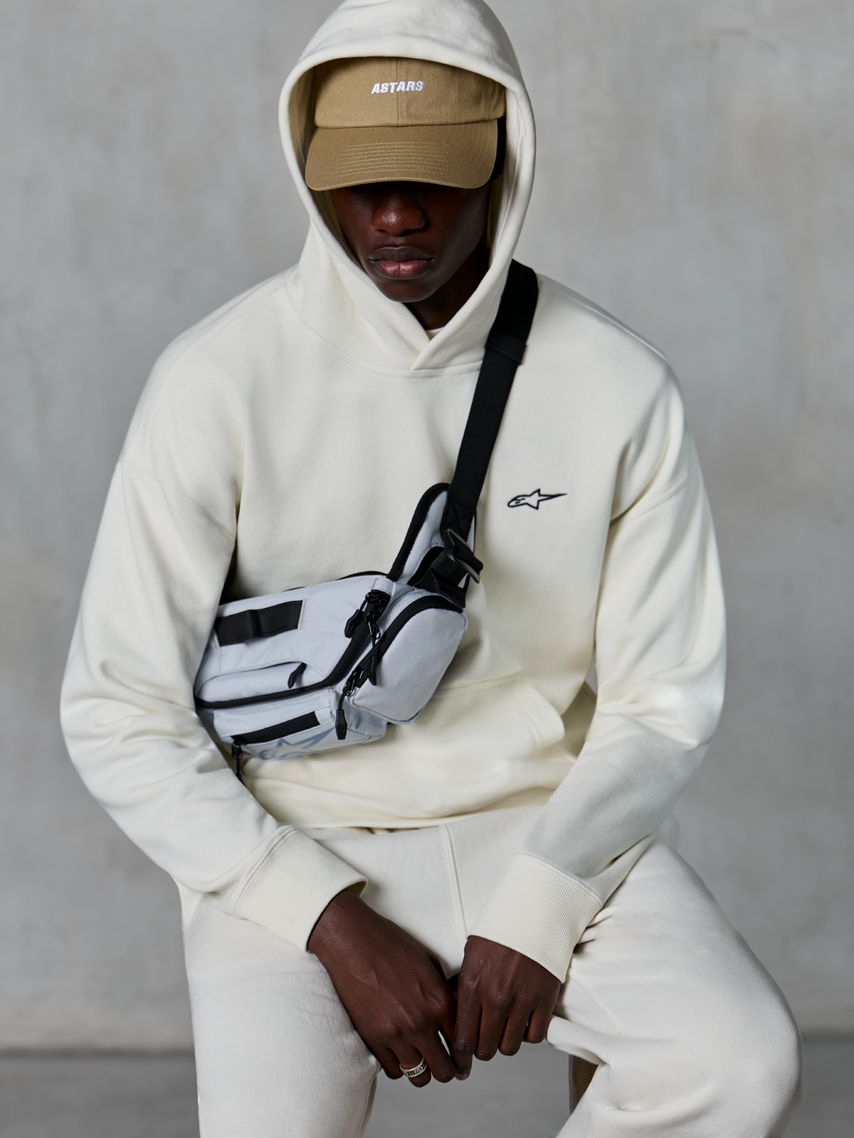 Alpinestars Essentials Ageless Hoodie, streetwear casual, cor Off White, pequeno logótipo preto bordado no peito, capuz usado sobre um boné bege com a marca ASTARS, combinado com uma mala de tiracolo cinzenta clara, vestuário lifestyle premium