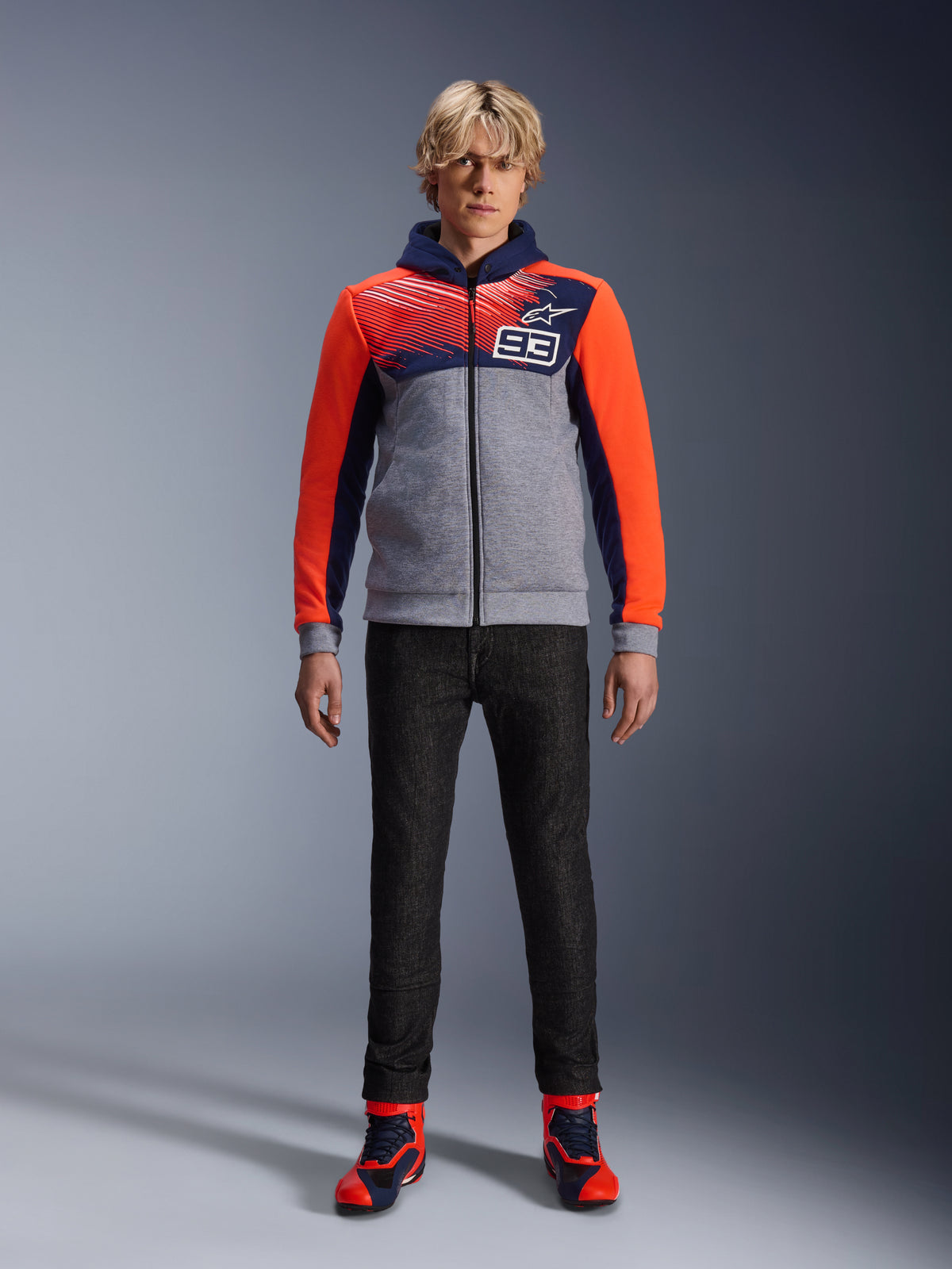 Alpinestars MM93 Chrome V2 Sport Hoodie, hoodie de motociclismo, azul marinho, cinzento mesclado e vermelho fluo, usado com calças de condução escuras e sapatilhas com detalhes vermelhos, com fecho de correr integral e logótipo MM93, design casual urbano inspirado no desporto motorizado.