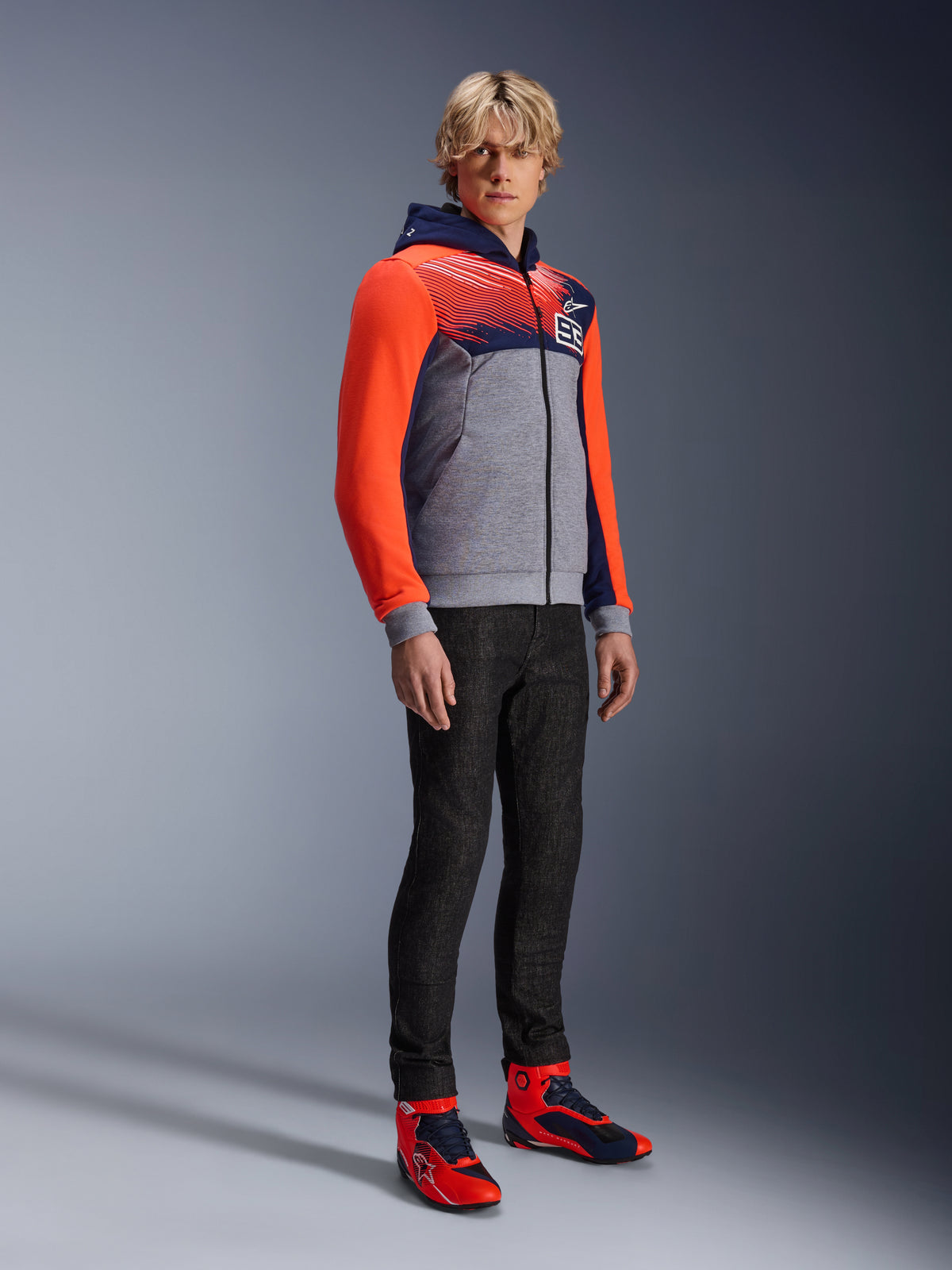 Alpinestars MM93 Chrome V2 Sport Hoodie, hoodie de motociclismo urbano, corpo cinzento mesclado com peito azul-marinho e mangas vermelho néon brilhante, com o logótipo 93 no peito, usado por um modelo com calças pretas de corte justo e sapatilhas de condução de cano alto coordenadas em vermelho e azul, corte atlético moderno para condução urbana