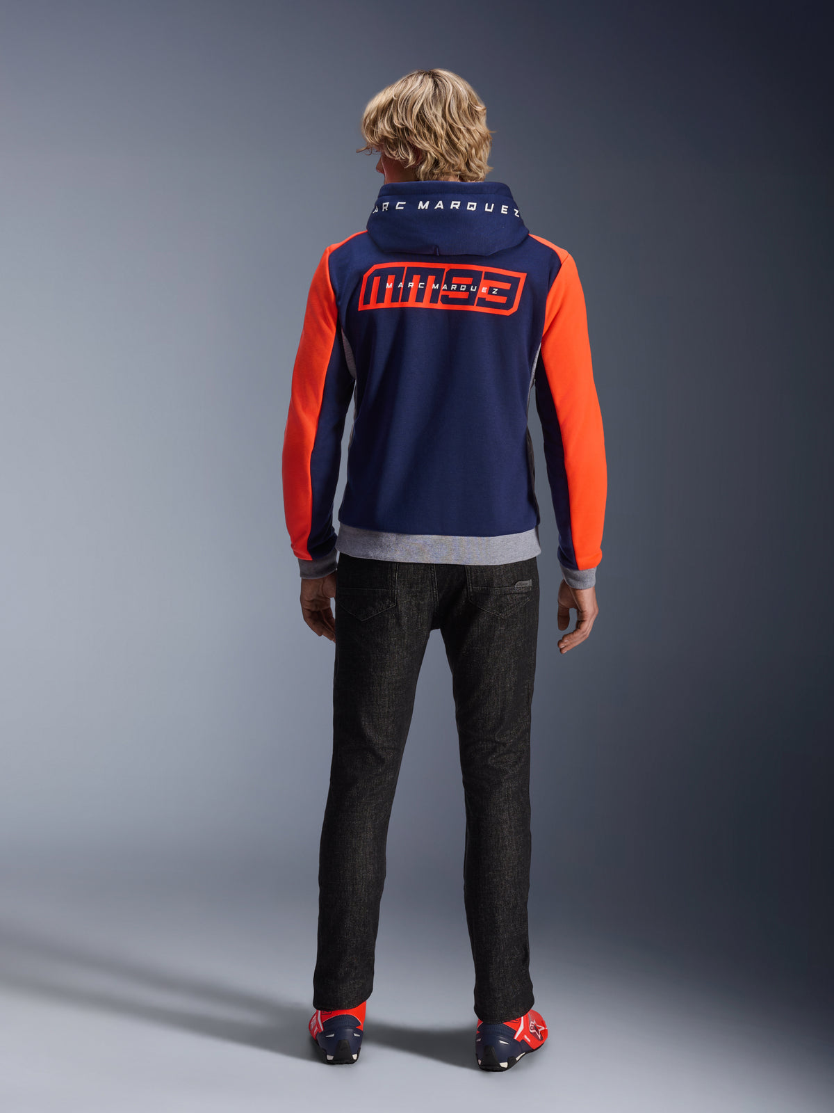 Modelo de costas a usar o Alpinestars MM93 Chrome V2 Sport Hoodie, azul marinho com mangas vermelho fluo e detalhes em cinzento mesclado, grandes logótipos MM93 e Marc Marquez nas costas e no capuz, hoodie casual de motociclismo combinado com calças pretas