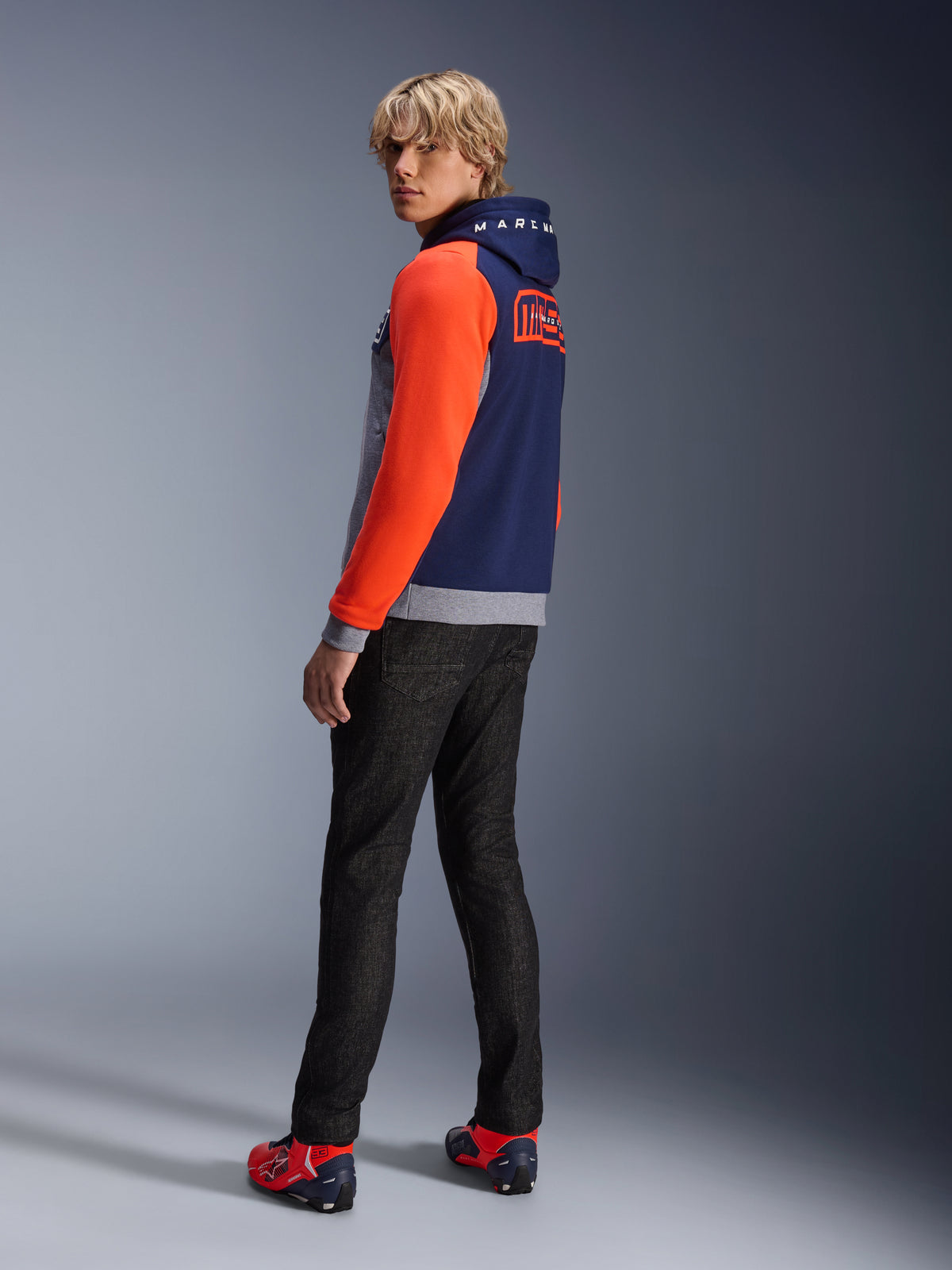 Hoodie Alpinestars MM93 Chrome V2 Sport, Azul Marinho, Cinza Melange e Vermelho Fluo, modelo a usar hoodie com mangas em vermelho vivo e logótipo MM93 Marc Marquez nas costas, combinado com calças de ganga escuras e sapatos de condução vermelhos, equipamento de motociclismo urbano casual