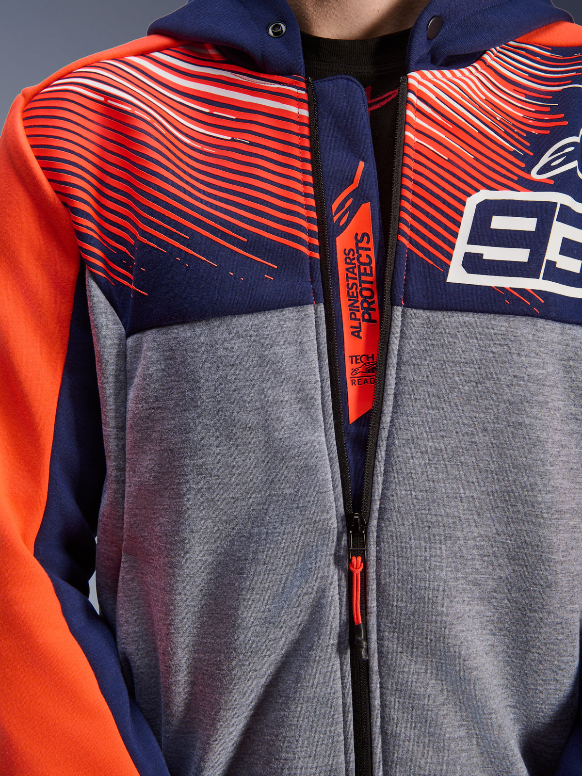 Alpinestars MM93 Chrome V2 Sport Hoodie, pormenor do peito e colarinho, tecido polar técnico azul marinho e cinzento melange com gráficos vermelho fluo, logótipo 93 branco e marca Alpinestars Protects na aba interna do fecho de correr, vestuário de motociclismo Tech-Air Ready