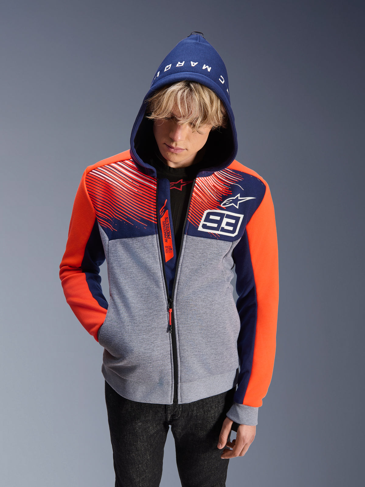 Alpinestars MM93 Chrome V2 Sport Hoodie, hoodie de motociclismo, corpo cinzento mesclado com parte superior azul-marinho e mangas cor de laranja fluorescente, com o número 93 branco e o logótipo da estrela Alpinestars, texto Marc Marquez no capuz, usado por um modelo masculino com o capuz colocado, design de inspiração motorsport urbana