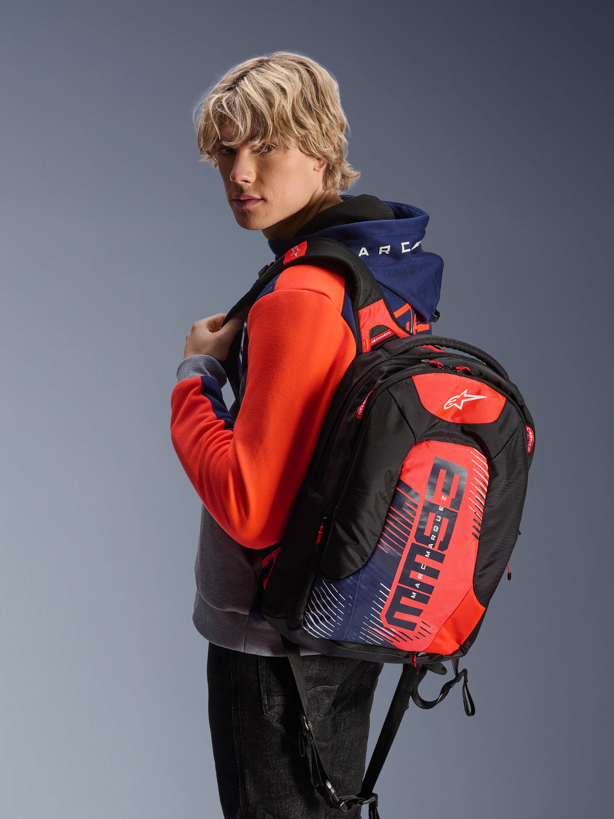 Modelo masculino com o Alpinestars MM93 Chrome V2 Sport Hoodie em azul marinho, cinzento mesclado e vermelho néon, combinado com uma mochila MM93 a condizer, vestuário casual de motociclismo urbano com uma silhueta atlética moderna