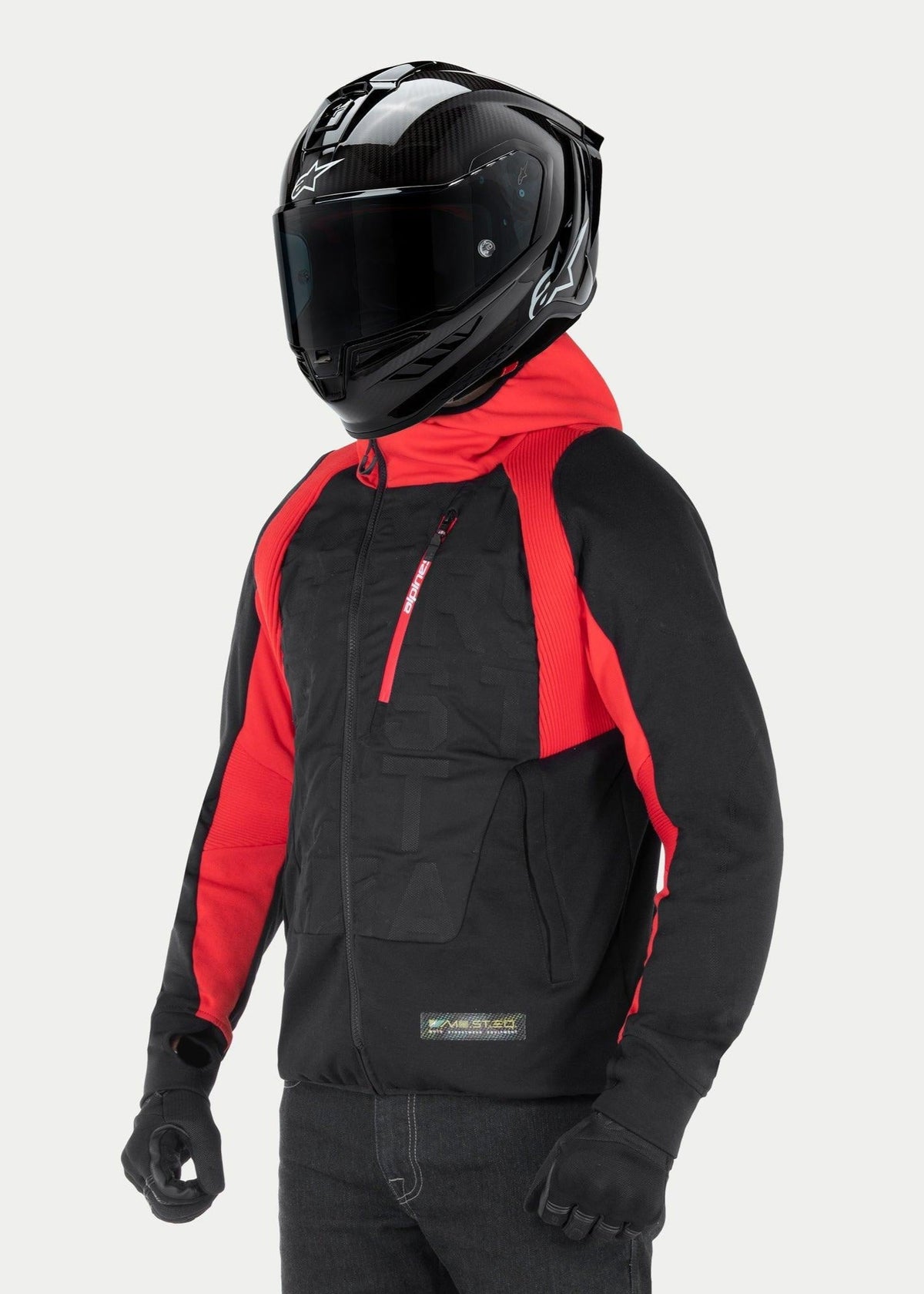 Casaco com Capuz Híbrido Alpinestars MO.ST.EQ, hoodie de motociclismo urbano, preto e vermelho, com um fecho de correr frontal inclinado e painéis canelados nos ombros, usado com capacete integral preto e luvas pretas, estilo streetwear contemporâneo