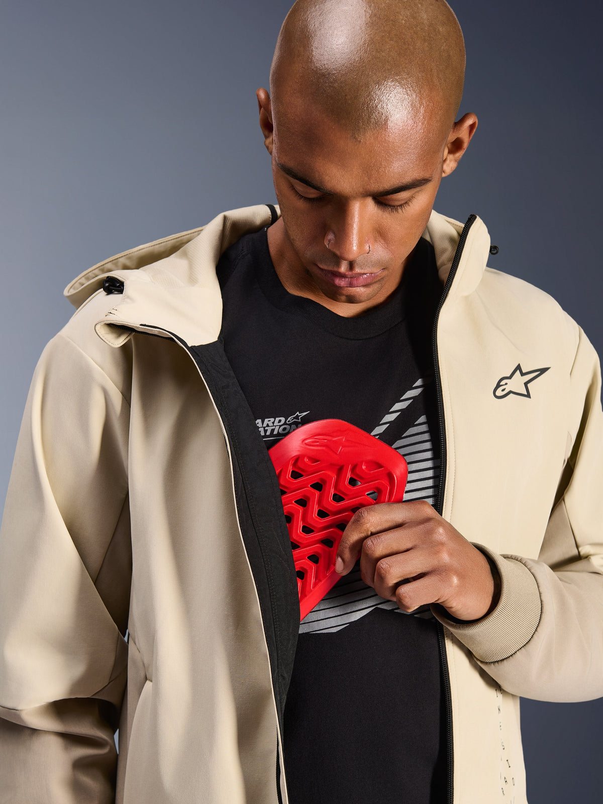 Alpinestars Radium Tech Hoodie em Mastic Aluminum, casaco de motociclo urbano, modelo a demonstrar o bolso interno para armadura com um protetor de peito Nucleon vermelho, estrutura técnica corta-vento com um design casual de hoodie e logótipo Alpinestars.