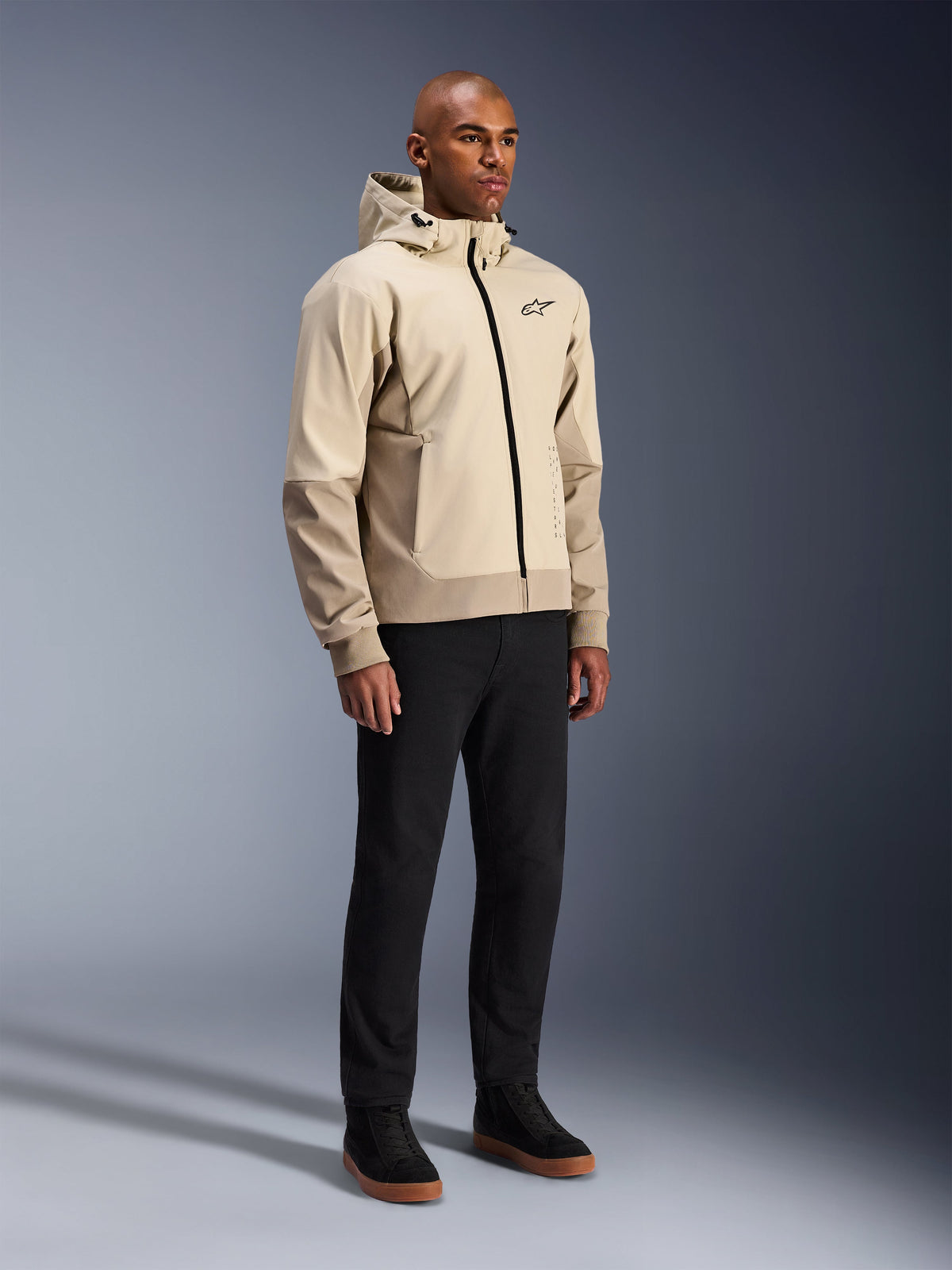 Alpinestars Radium Tech Hoodie, casaco de motociclismo urbano, bege claro Mastic Aluminum, usado com calças pretas e sapatilhas de cano alto pretas, com uma camada técnica corta-vento, fecho frontal e capuz amovível, design streetwear moderno para condução urbana