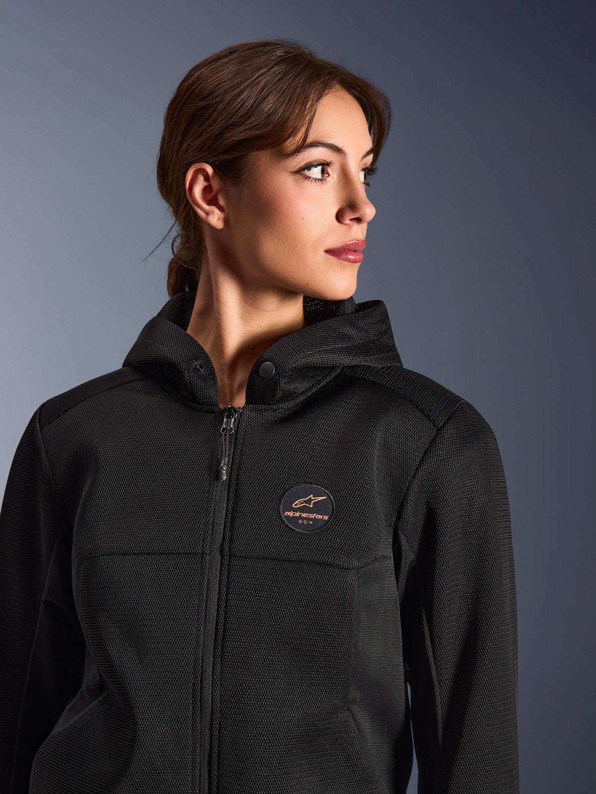 Mulher a usar o hoodie Alpinestars Stella Chrome Superairflow, preto com logótipo em cobre, construção em malha 3D para máximo fluxo de ar, equipamento técnico de motociclismo concebido para mulheres, estilo de condução desportiva urbana