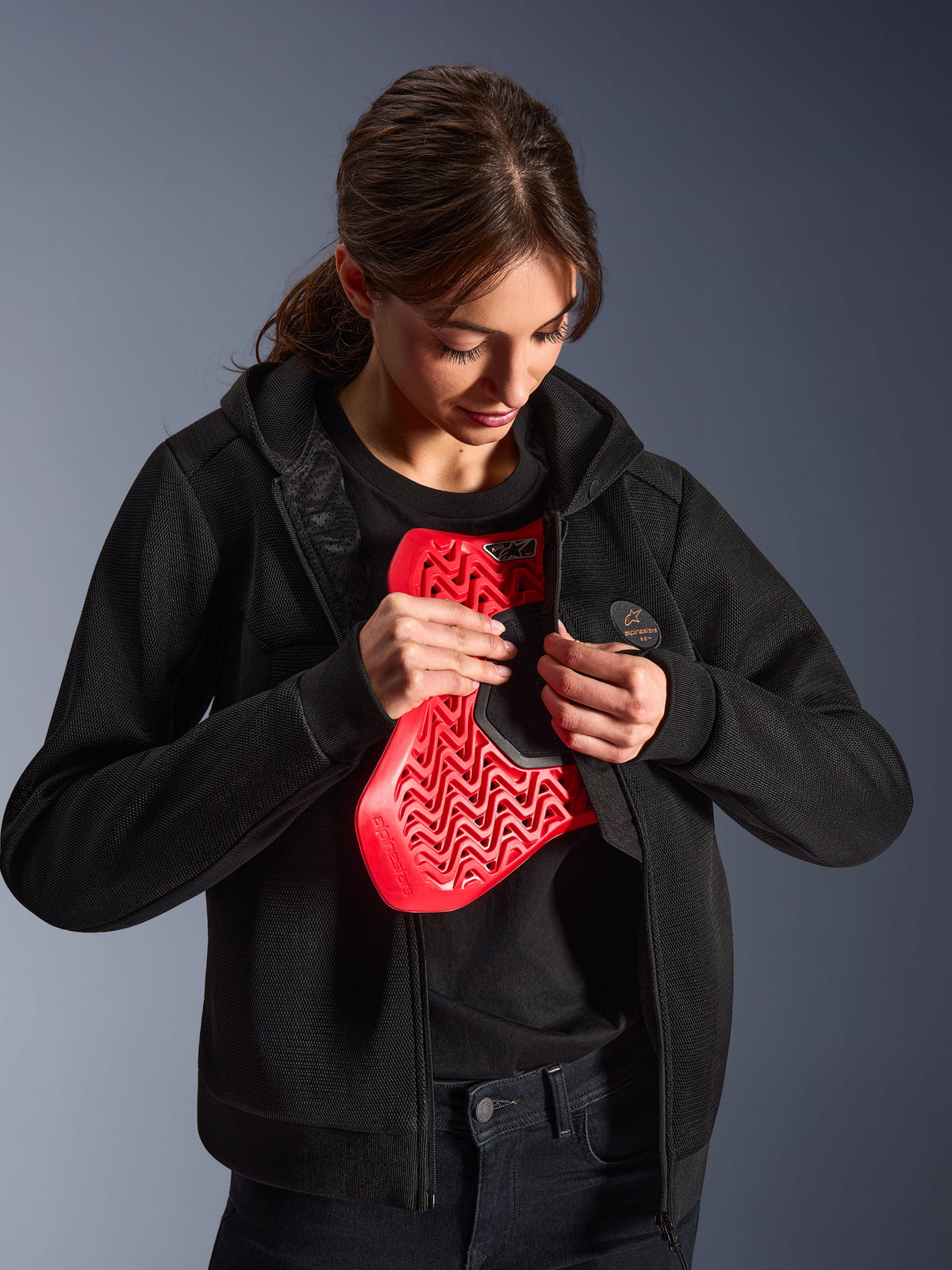 Alpinestars Stella Chrome Superairflow Hoodie, sweatshirt com capuz de motociclismo para mulher, preta, modelo a segurar um protetor de peito Nucleon vermelho, construção em malha 3D leve para condução urbana e desportiva