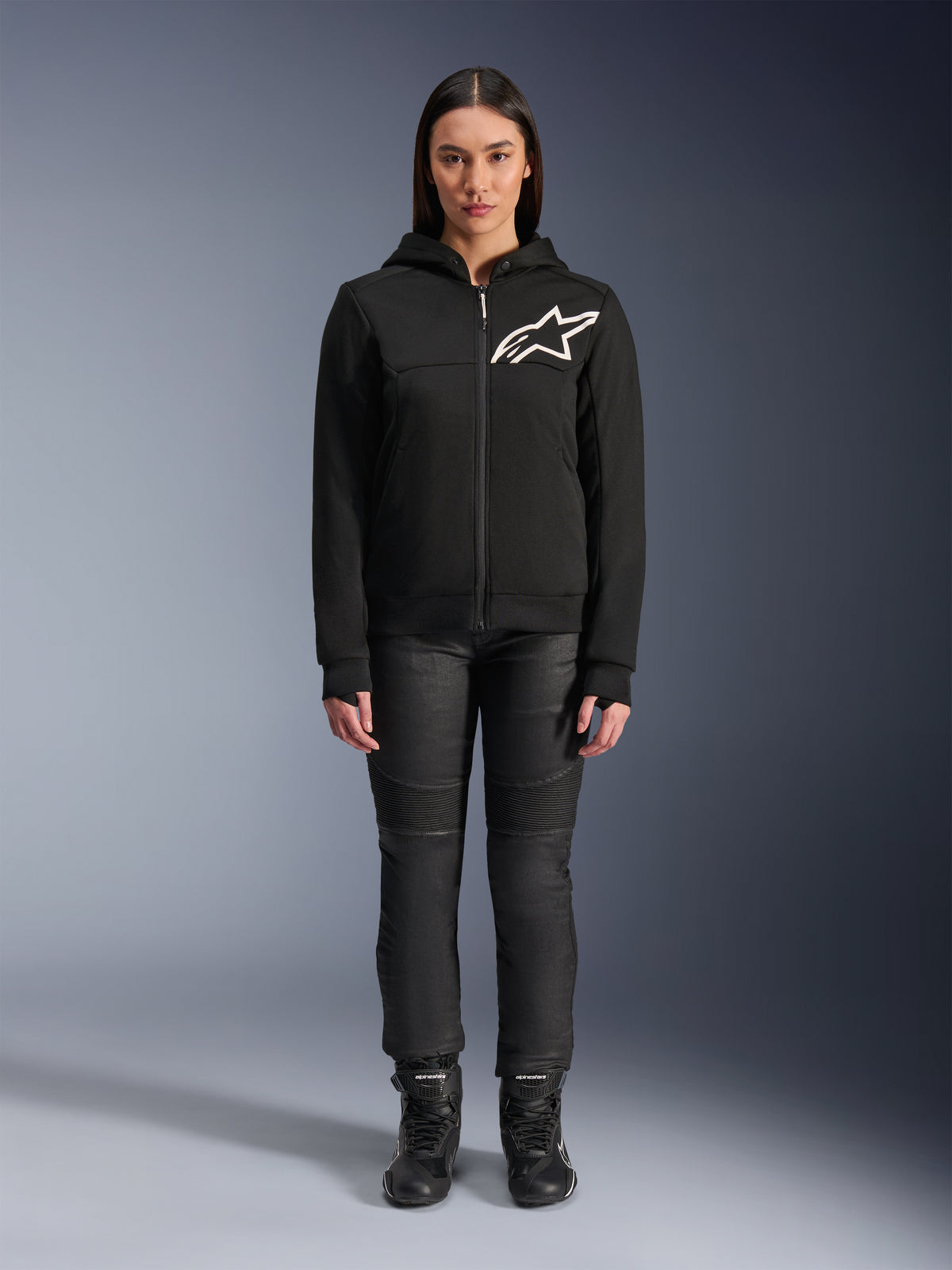 Mulher a usar o Alpinestars Stella Chrome V2 Sport Hoodie, preto com logótipo Astars branco no peito, combinado com calças técnicas de condução pretas e botas, design de vestuário urbano para motociclismo