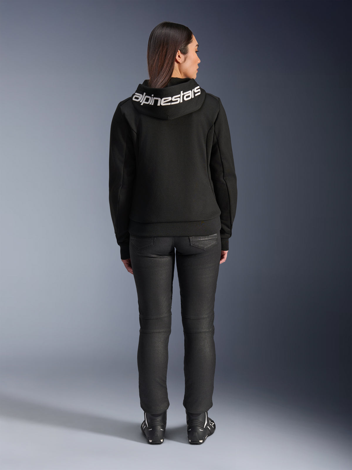 Vista traseira de uma modelo a usar o Hoodie Alpinestars Stella Chrome V2 Sport em preto, com um logótipo Alpinestars branco proeminente no capuz, combinado com calças de motociclismo pretas e botas de mota, equipamento de motociclismo desportivo urbano para mulher