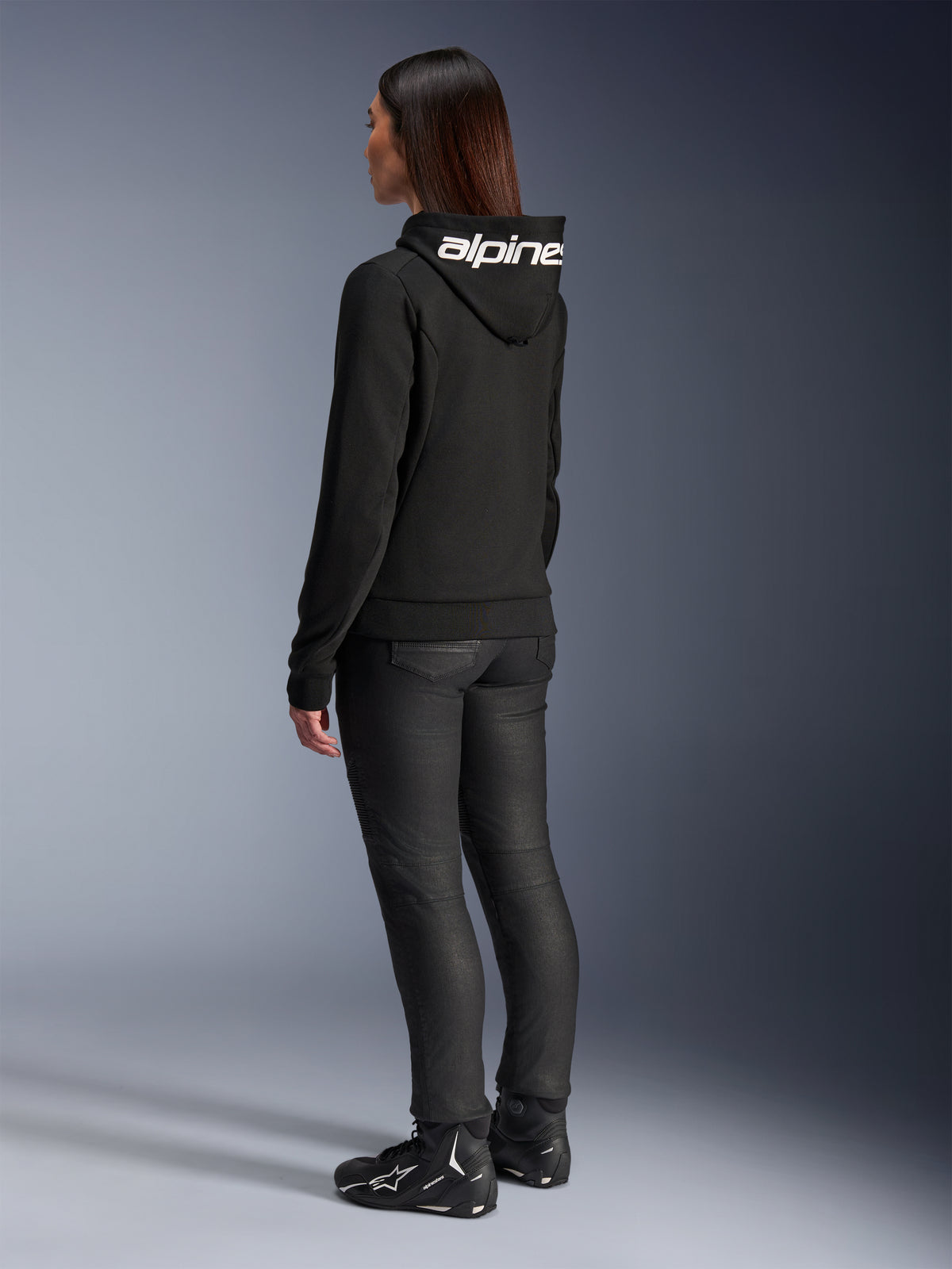 Mulher com o Hoodie Alpinestars Stella Chrome V2 Sport, vista traseira, hoodie de motociclismo preto com logótipo Alpinestars branco no capuz, combinado com calças técnicas de condução pretas e calçado de motociclismo preto, equipamento casual para deslocações urbanas