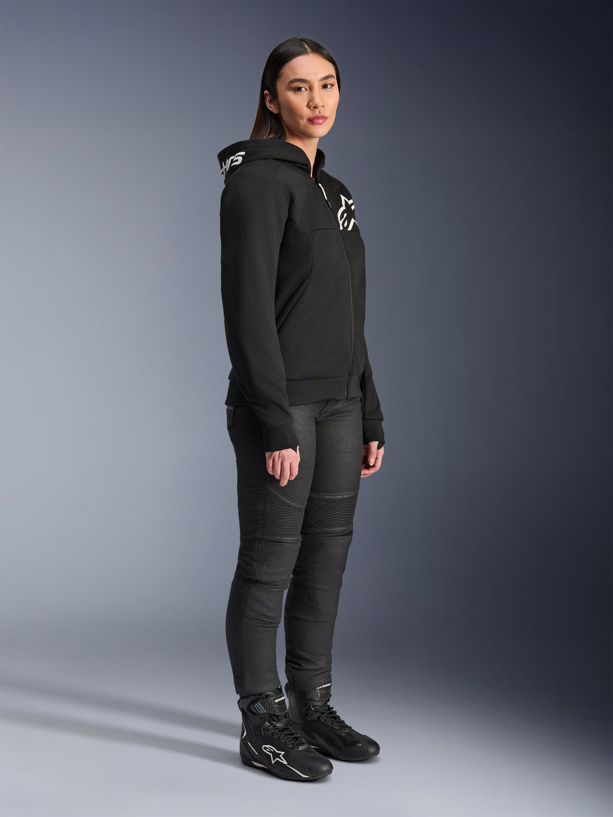 Mulher a usar Hoodie Alpinestars Stella Chrome V2 Sport em preto, com logótipo Alpinestars em branco, combinado com calças e botas de motociclismo pretas, equipamento de condução urbana para mulher com um ajuste atlético casual