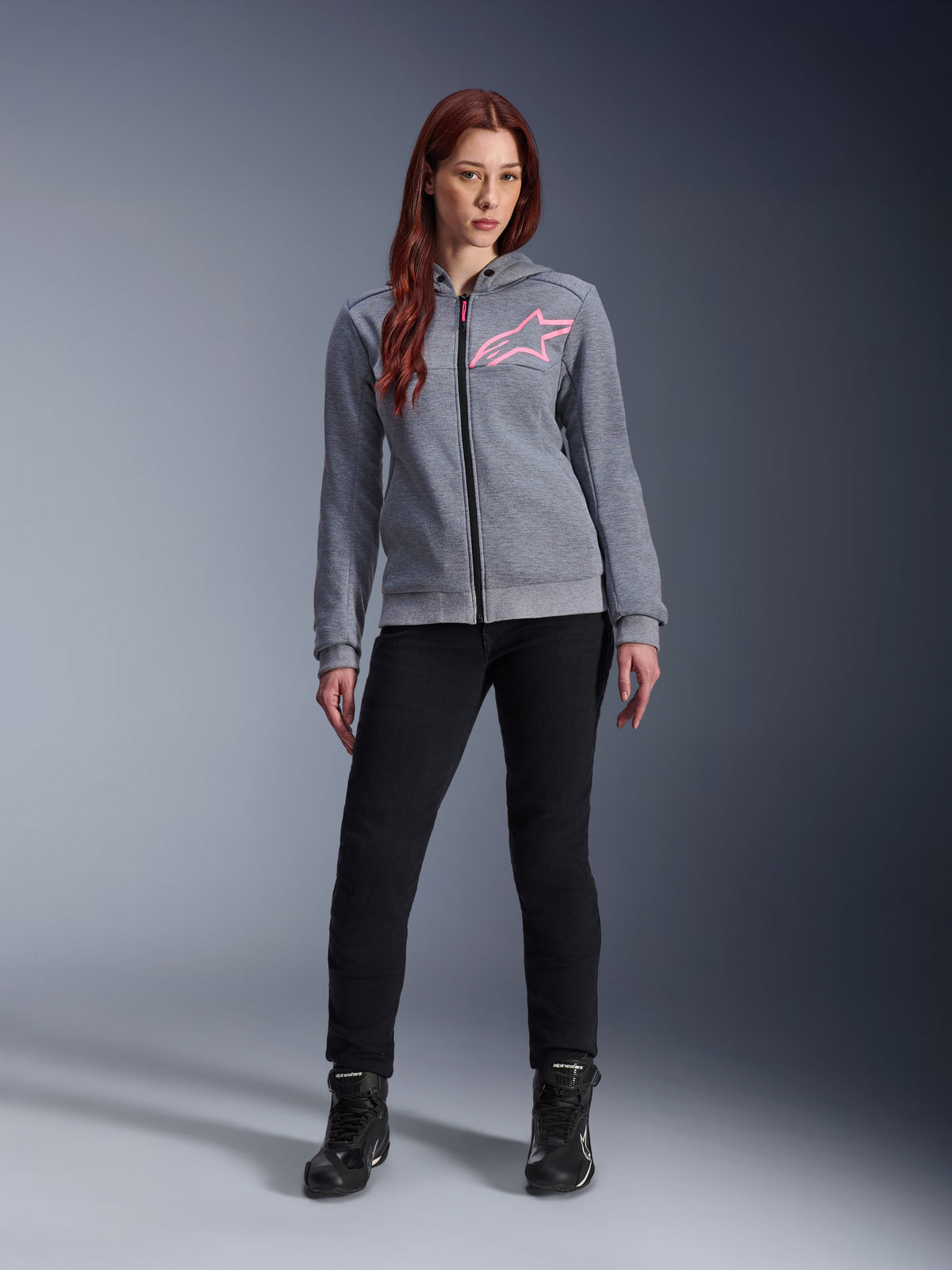 Alpinestars Stella Chrome V2 Sport Hoodie, cinzento mesclado e rosa fluorescente, usado por uma mulher com calças e botas pretas, com o logótipo de estrela Alpinestars rosa no peito, equipamento de motociclismo urbano com fecho integral, polar técnico leve com um ajuste atlético casual.