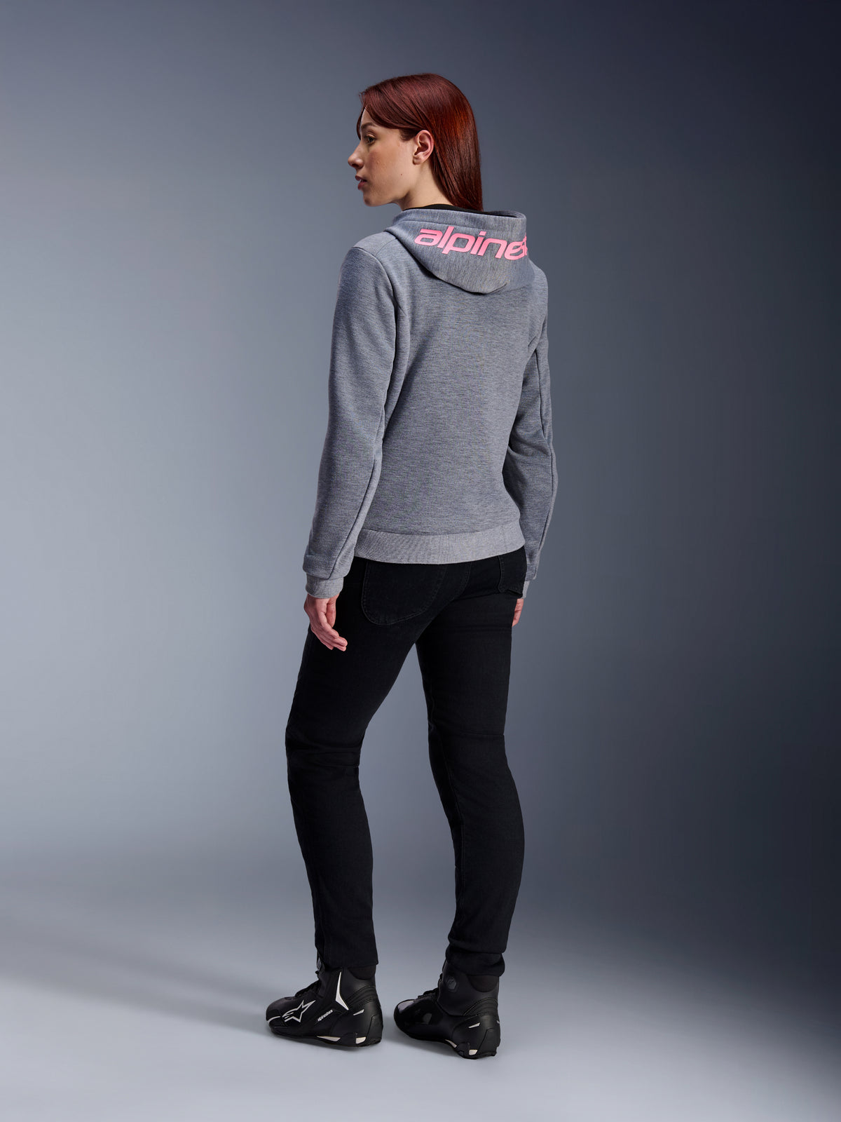 Alpinestars Stella Chrome V2 Sport Hoodie, hoodie de motociclismo para mulher, cinza mesclado com logótipo rosa fluorescente no capuz, vista de costas, usado com calças pretas e calçado de condução, estilo urbano casual com proteção para motociclismo
