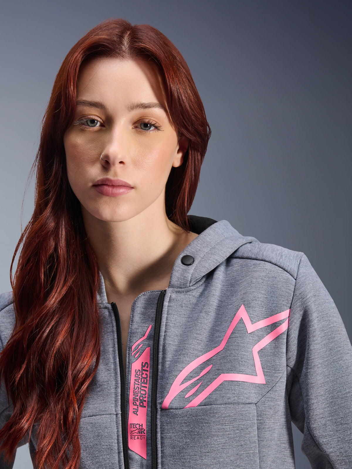 Mulher a usar o Alpinestars Stella Chrome V2 Sport Hoodie, Gray Heather Pink Fluo, grande plano que mostra o logótipo de estrela cor-de-rosa e a marca Tech-Air Ready, vestuário urbano para motociclismo.