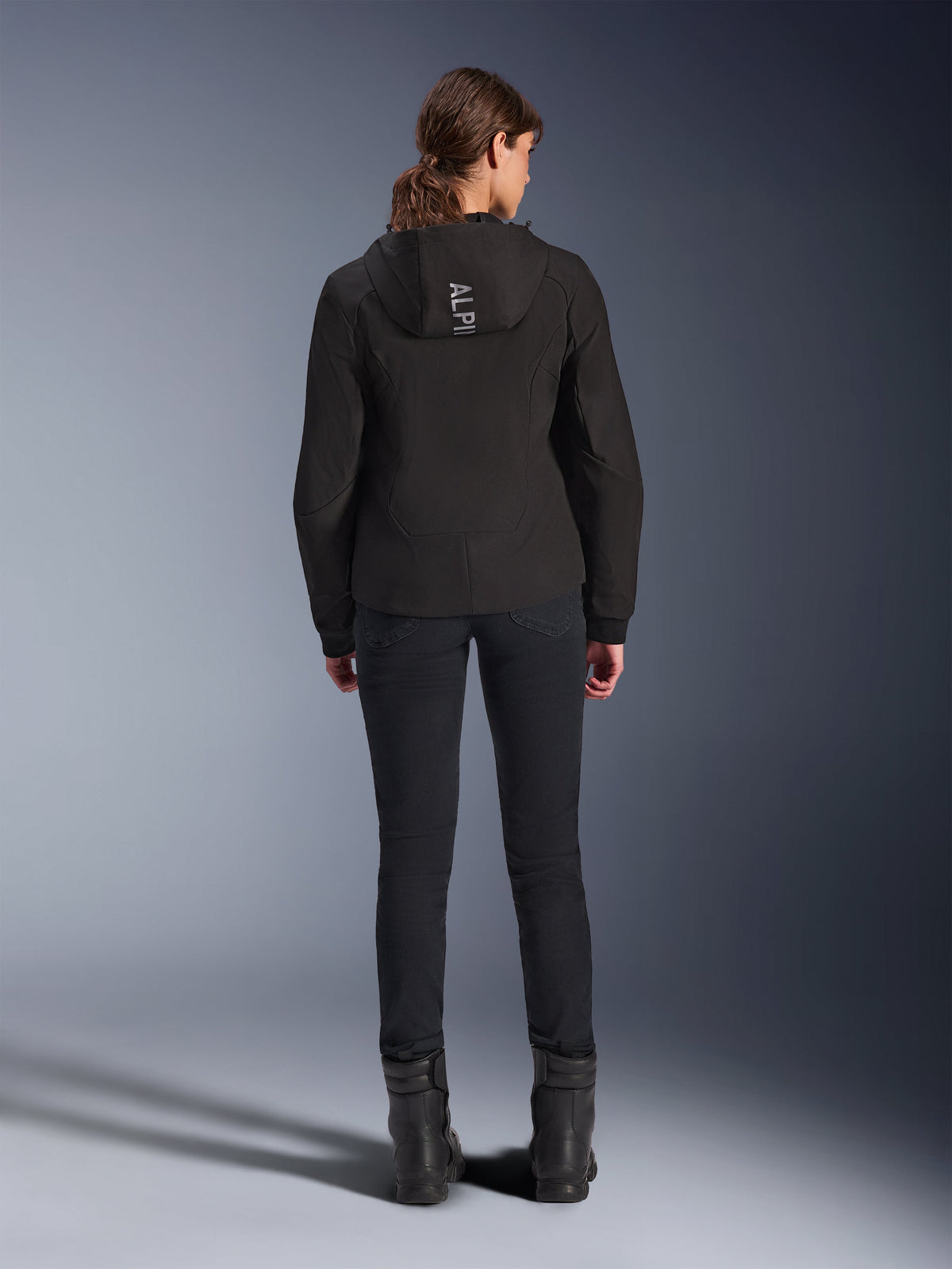 Alpinestars Woman Stella Radium Tech Hoodie, preto, vista traseira de modelo a usar hoodie técnico leve de 3 camadas resistente ao vento com logótipo Alpinestars no capuz, combinada com calças e botas pretas, vestuário de condução urbana em motociclo