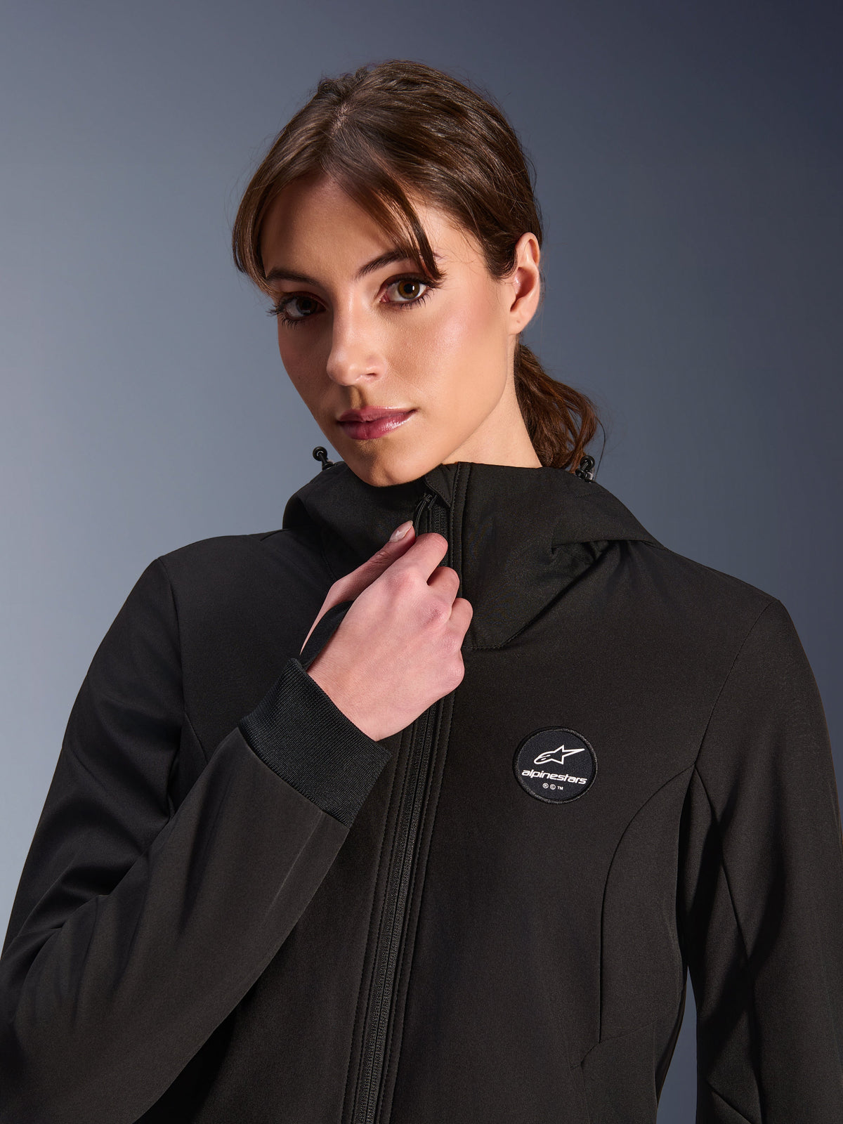 Hoodie Stella Radium Tech para Mulher, preto, vestido por uma mulher a ajustar o fecho frontal, com logótipo circular da Alpinestars no peito, punhos elásticos canelados e construção têxtil corta-vento para condução urbana