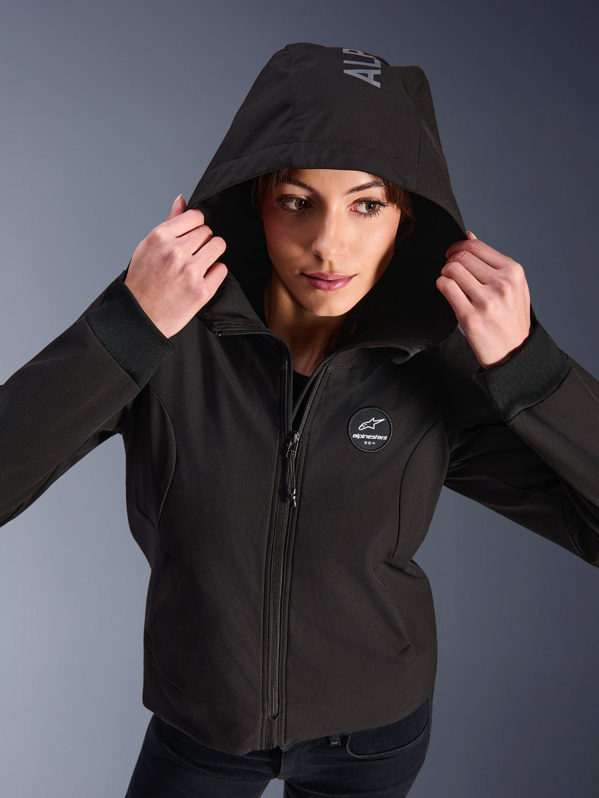 Alpinestars Stella Radium Tech Hoodie em preto, mulher a usar hoodie de motociclismo urbano com o capuz levantado, logótipo circular da Alpinestars no peito, punhos canelados e construção de 3 camadas corta-vento