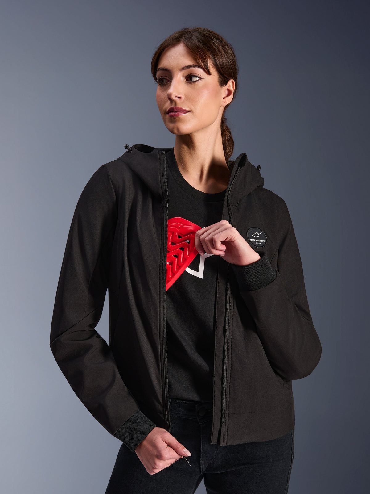 Alpinestars Woman Stella Radium Tech Hoodie, hoodie preta de condução urbana, modelo feminina a demonstrar a armadura vermelha Nucleon Flex Plus no interior, estrutura têxtil leve de 3 camadas resistente ao vento com punhos canelados e logótipo no peito, vestuário de proteção para motociclismo