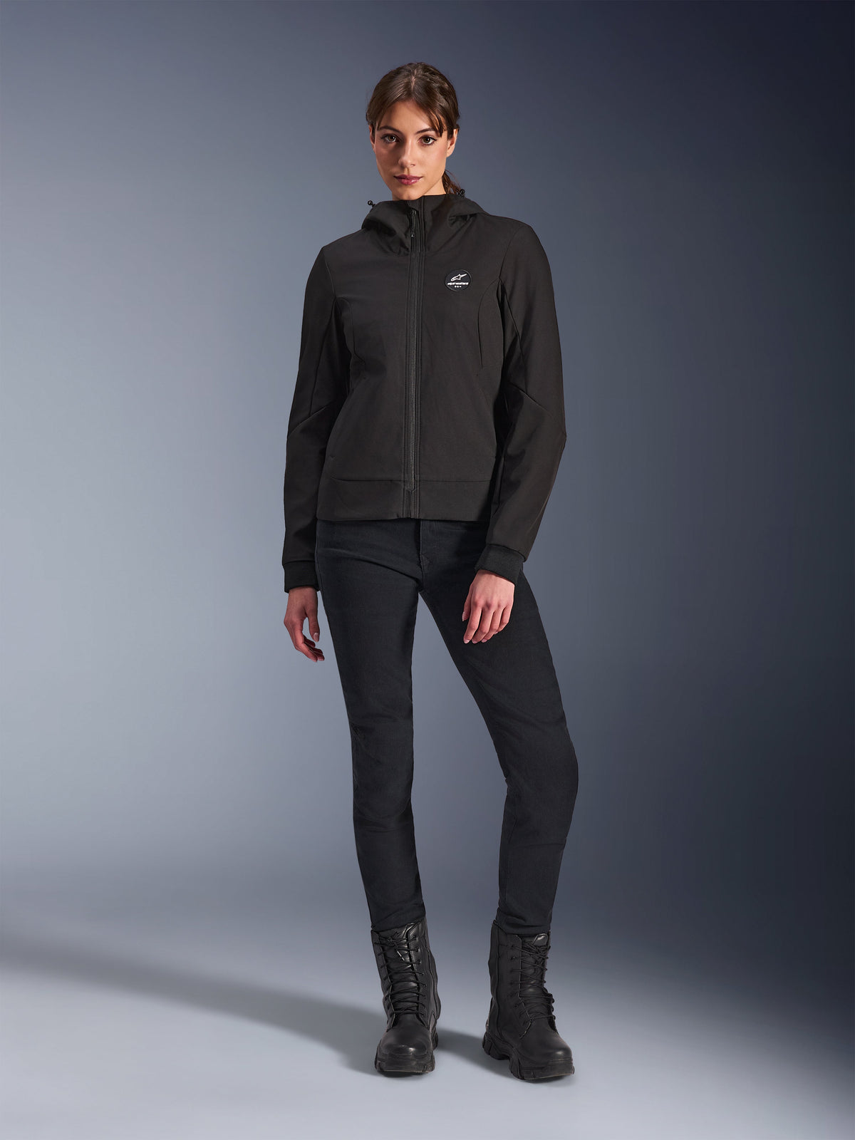 Alpinestars Stella Radium Tech Hoodie em preto, camisola técnica de motociclismo urbana para mulher, usada com calças pretas de corte justo e botas pretas, apresentando um pequeno logótipo no peito e capuz, construção leve corta-vento para condução urbana
