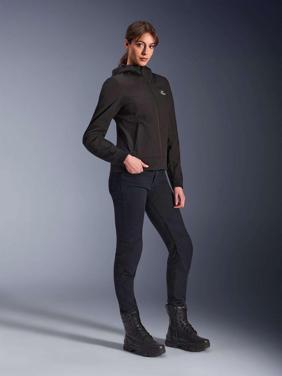 Alpinestars Stella Radium Tech Hoodie, hoodie de motociclismo urbana preta para mulher, usada com calças de condução pretas e botas de estilo tático, corte atlético justo, estrutura técnica de 3 camadas corta-vento, design minimalista com pequeno logótipo no peito, fotografia de estúdio de corpo inteiro
