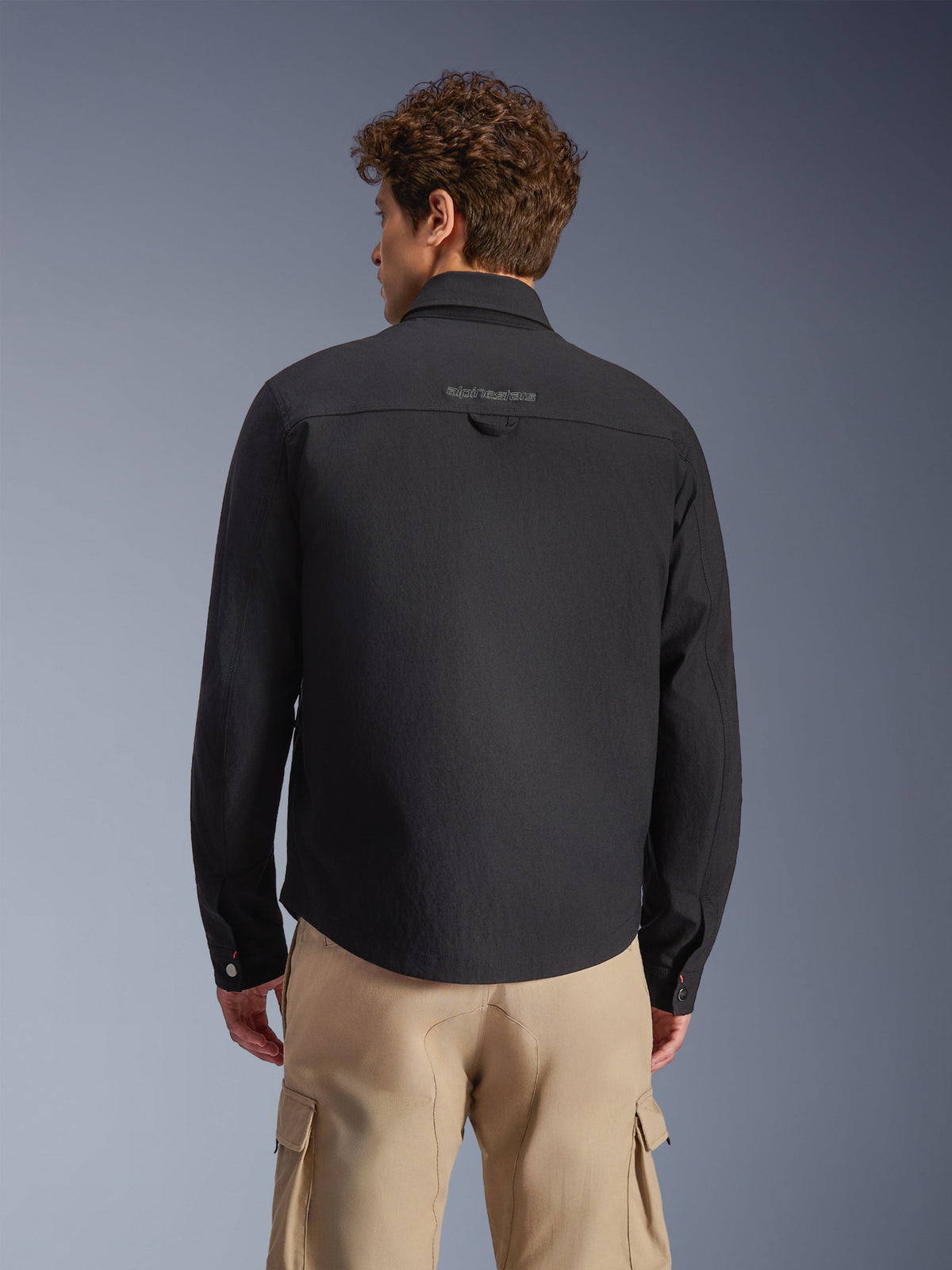Sobrecamisa Alpinestars Bryon Resistente à Água, preta, vista traseira num modelo, com marca bordada na pala traseira e alça utilitária, combinada com calças cargo bege, equipamento de motociclismo urbano com estética clássica de roupa de trabalho