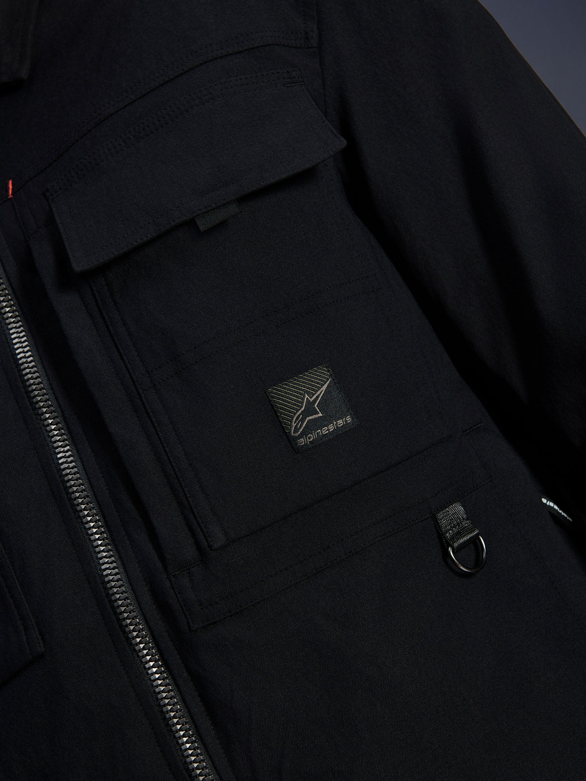 Alpinestars Bryon Water Resistant Overshirt, preta, vista de pormenor do tecido de lona encerada, fecho de correr frontal, bolso cargo no peito com emblema do logótipo Alpinestars e detalhe de anel em D metálico