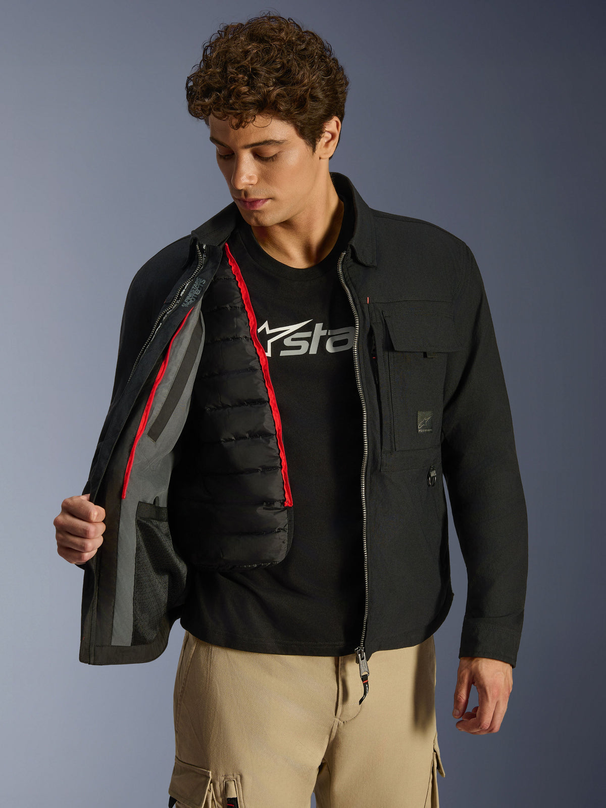 Alpinestars Bryon WR Overshirt, preto, casaco de motociclismo urbano mostrado aberto para revelar um colete térmico acolchoado preto amovível com debrum vermelho, usado com calças cargo bege e uma t-shirt com logótipo, design funcional inspirado em vestuário de trabalho