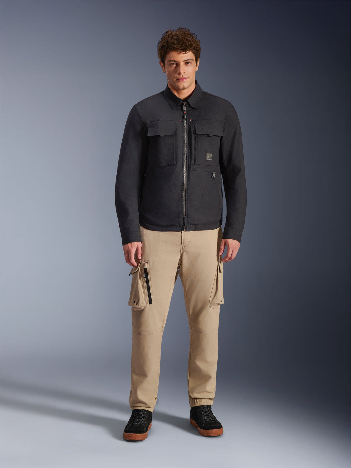 Modelo masculino a usar a sobrecamisa Alpinestars Bryon Water Resistant em preto, com bolsos cargo no peito e fecho central, combinada com calças cargo bege e sapatilhas pretas, retrato de estúdio de corpo inteiro.