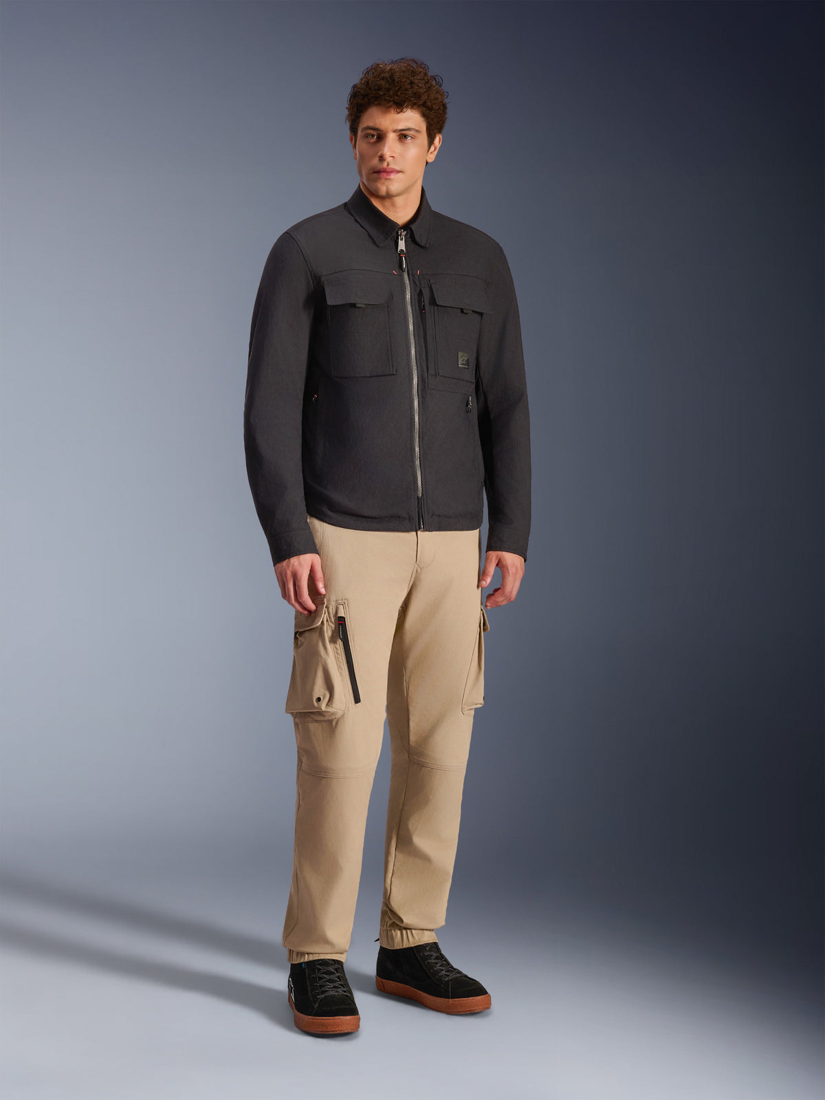 Sobrecamisa Alpinestars Bryon Water Resistant, preta, usada por modelo masculino com calças cargo caqui e sapatos pretos, casaco de motociclismo urbano com design inspirado em vestuário de trabalho, fotografia de corpo inteiro em estúdio