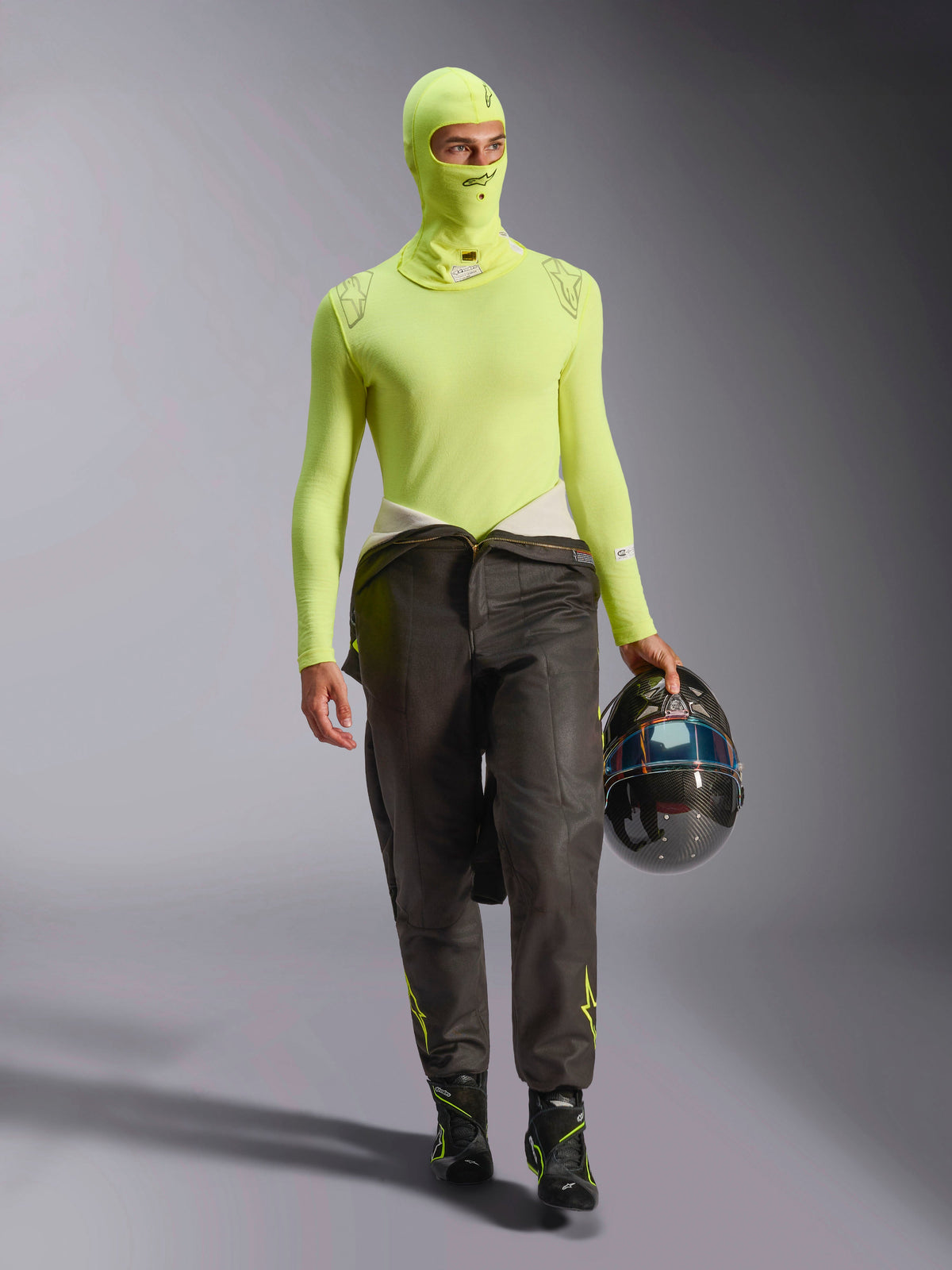 Piloto de corridas masculino a usar a Balaclava Alpinestars ZX Evo V3 em Yellow Fluo, camisola de base de manga comprida a condizer, calças de corrida escuras e botas de corrida pretas, a segurar um capacete de fibra de carbono, equipamento profissional de automobilismo