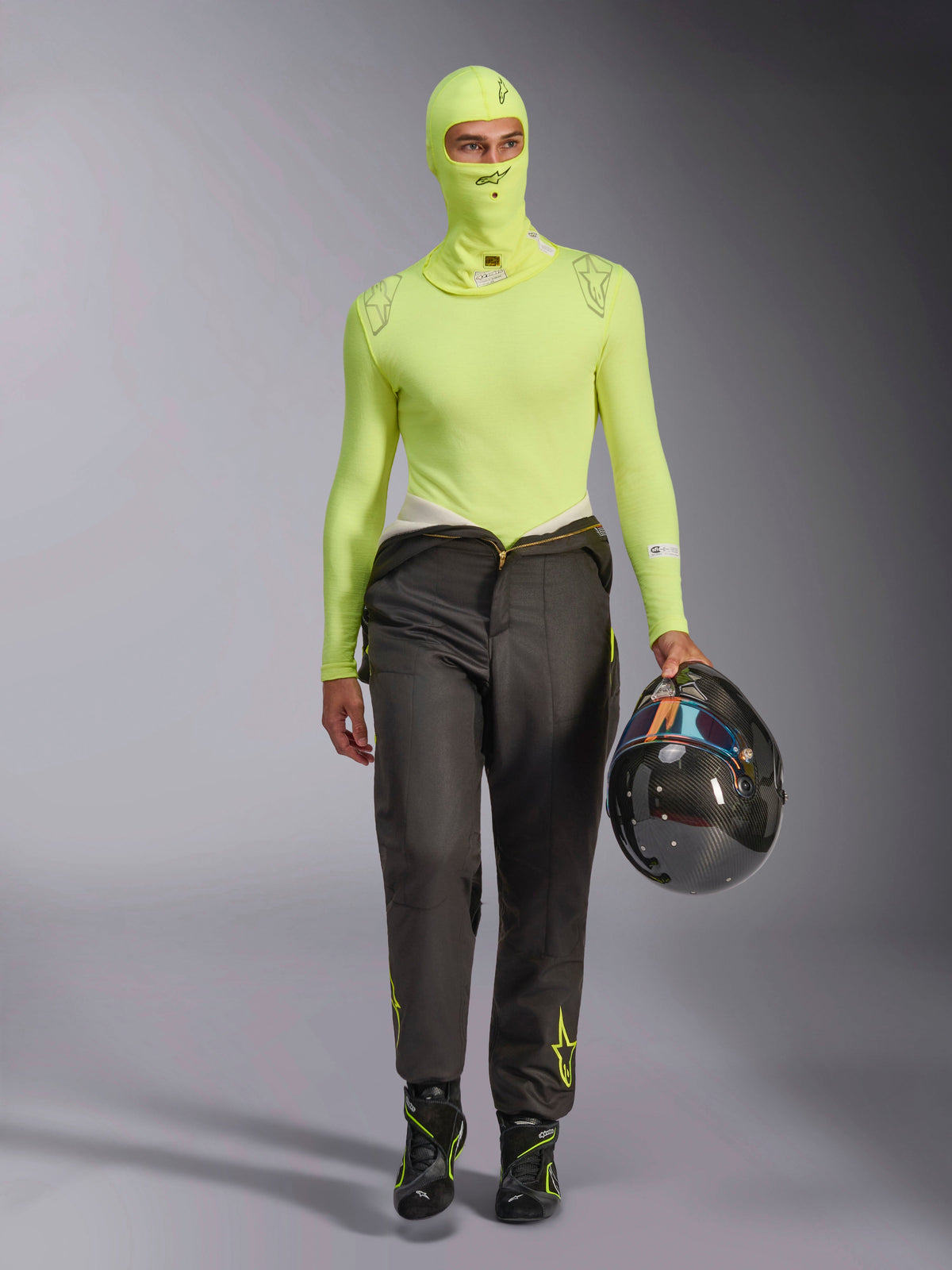 Balaclava Alpinestars ZX Evo V3 em amarelo fluo, usada com camisola de camada de base de manga comprida a condizer e calças de automobilismo cinzento escuro, modelo masculino a segurar um capacete de fibra de carbono, equipamento profissional de automobilismo