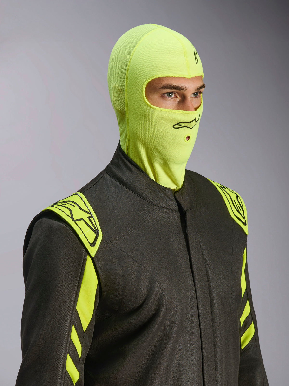 Modelo masculino a usar a balaclava Alpinestars ZX Evo V3 em Yellow Fluo, proteção de cabeça resistente ao fogo para competição, combinada com fato de automobilismo cinzento escuro com detalhes em amarelo néon, logótipo Alpinestars preto e abertura para tubo de hidratação, equipamento de proteção profissional para desportos motorizados