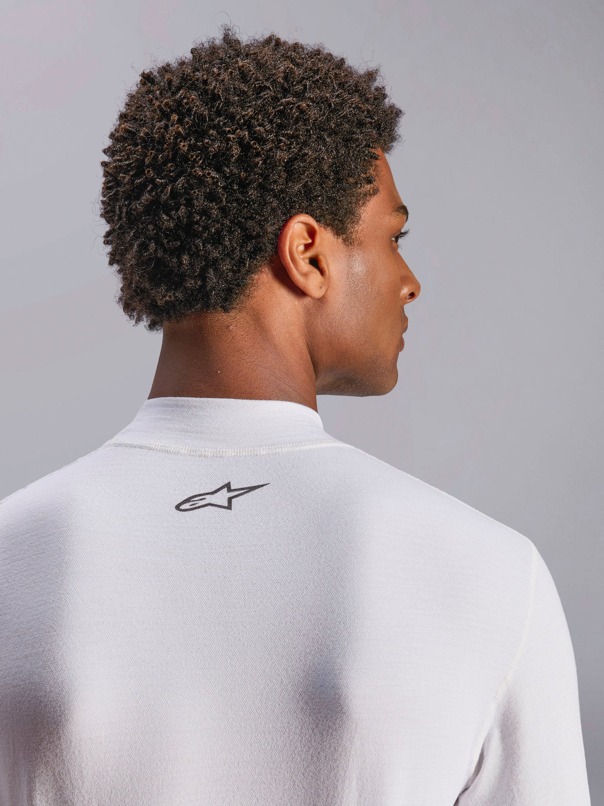 Alpinestars ZX Evo V3 Top, camada de base de manga comprida para automobilismo, branco, grande plano da zona da nuca com logótipo Alpinestars preto, tecido técnico resistente a chamas, usado por modelo com cabelo escuro encaracolado