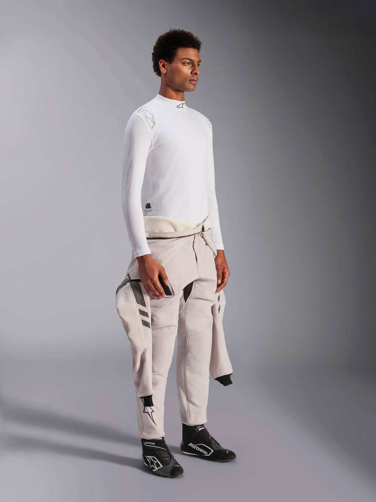 Alpinestars ZX Evo V3 Long Sleeve Top, camada interior de automobilismo branca resistente a chamas, vestida por um modelo com um fato de corrida atado à cintura, camada técnica leve com gola alta e detalhe do logótipo.