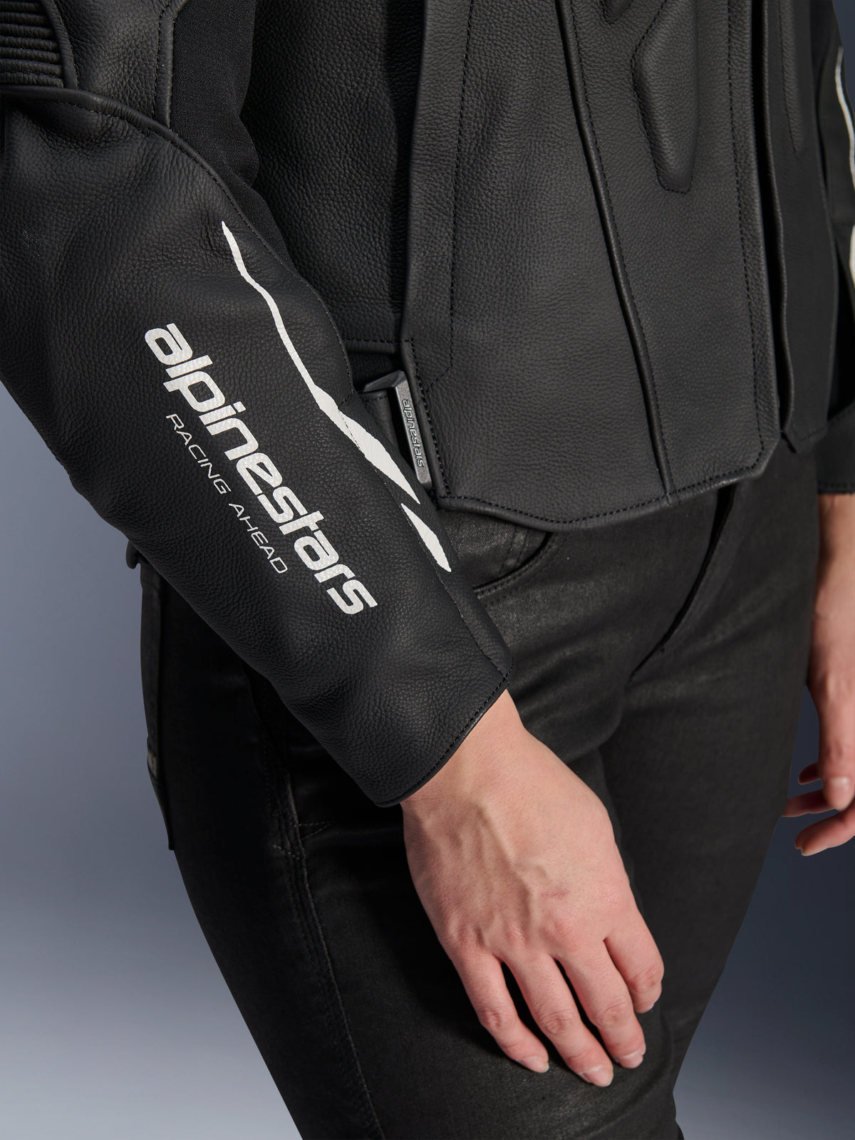 Pormenor do casaco de couro Stella Dusk da Alpinestars, casaco de motociclismo feminino em couro preto, com o logótipo branco 'alpinestars RACING AHEAD' na manga, textura de couro bovino premium, usado com calças pretas
