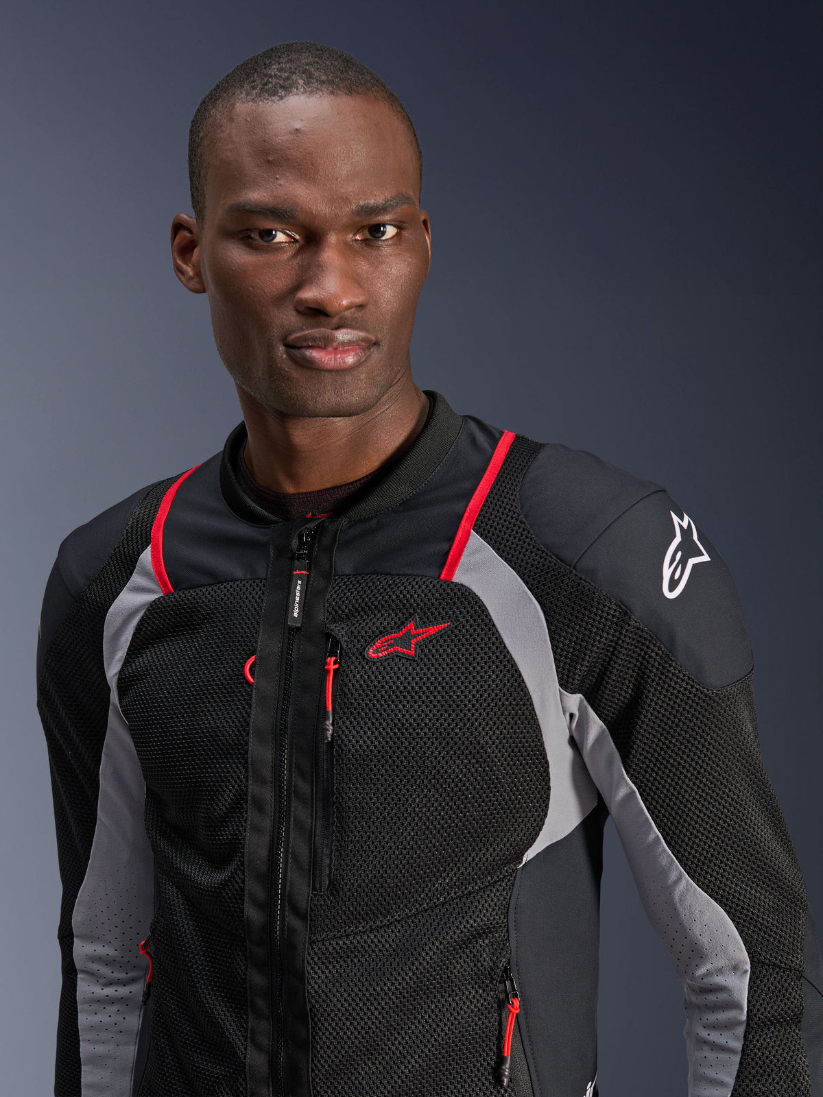 Homem a usar o Casaco Alpinestars Troop-Air Pro, preto, cinzento cinza e vermelho vivo, casaco de ar blindado técnico com painéis de malha respirável, detalhes em vivo vermelho e logótipo Alpinestars no ombro, equipamento de motociclismo urbano contemporâneo