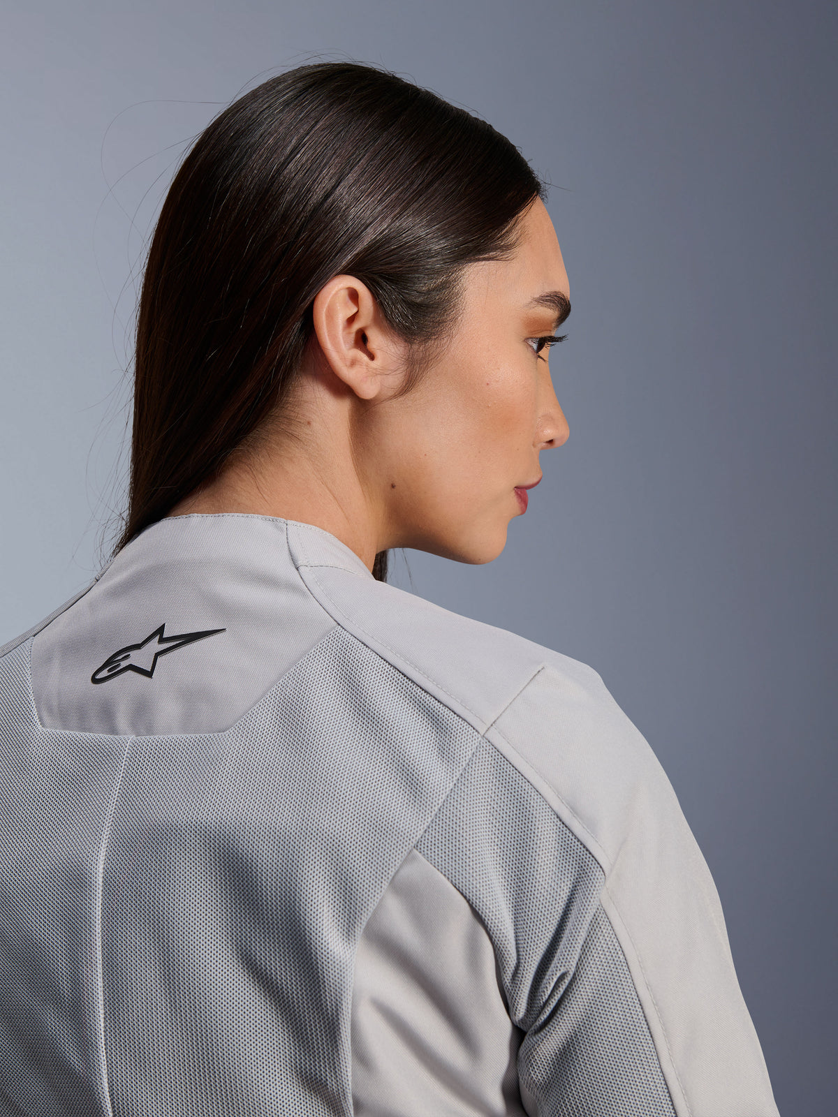Mulher a usar o casaco Alpinestars Stella C-1 Air em prateado, vista lateral-traseira em grande plano, mostrando o logótipo Alpinestars preto, painéis de ventilação em malha técnica e um design de motociclismo urbano contemporâneo e sóbrio.
