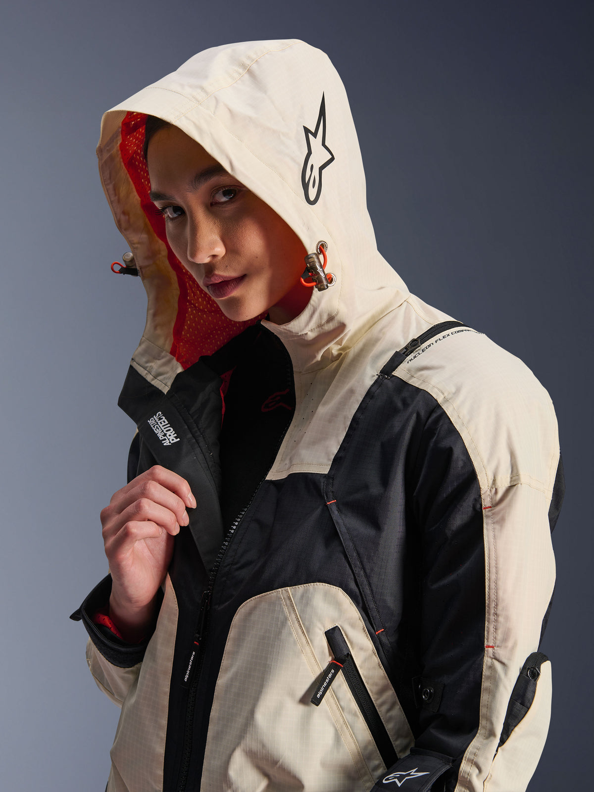 Casaco Alpinestars Molly Air em Black Almond, modelo feminina com o capuz colocado, com blocos de cor creme e preto, forro em malha laranja vibrante, elementos técnicos de design urbano, logótipo de estrela e detalhes de fechos