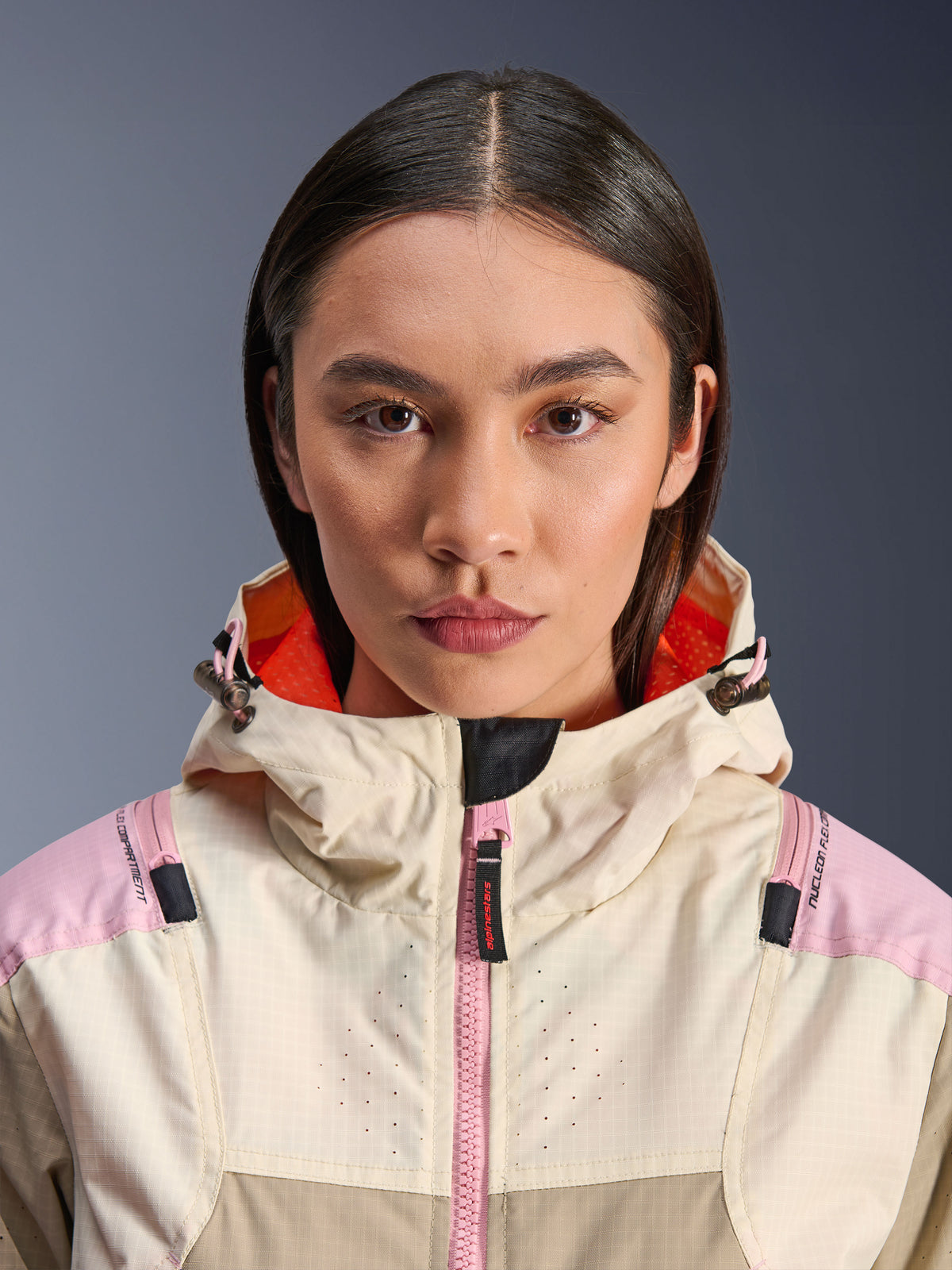 Primeiro plano de uma modelo feminina a usar o Casaco Alpinestars Molly Air em Sand Pink Almond, com gola alta e capuz com forro laranja vibrante, fecho frontal cor-de-rosa, tecido ripstop com peito perfurado a laser para ventilação, equipamento de proteção para motociclismo urbano
